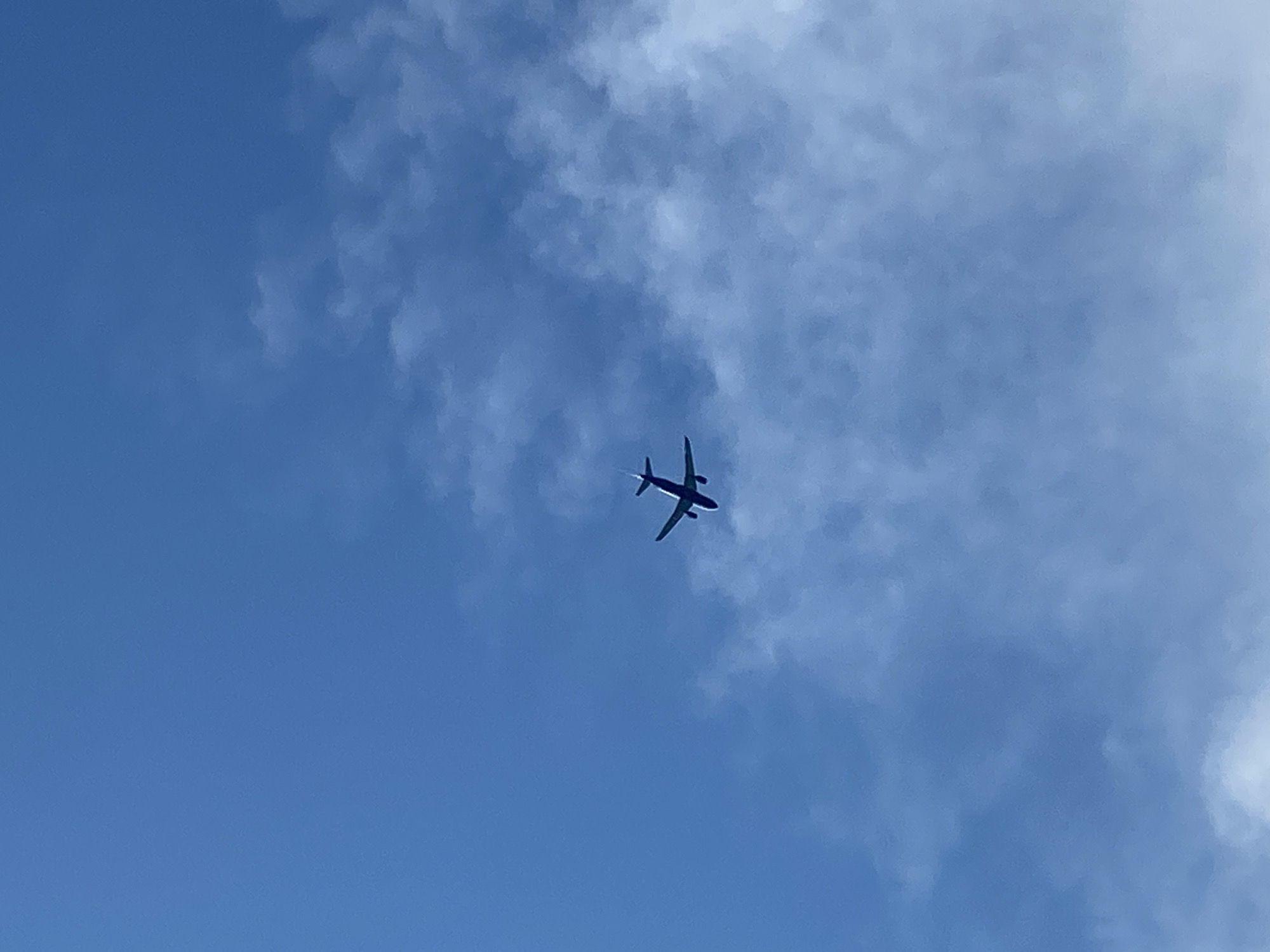 The image shows a blue sky with scattered clouds, suggesting daytime. A small airplane is visible, silhouetted against the sky, with its wings extended and a discernible tail. The airplane appears to be in mid-flight. The lighting suggests it is likely daytime. There is no visible text in the image. The context provided indicates the location is Prijevor, Montenegro.