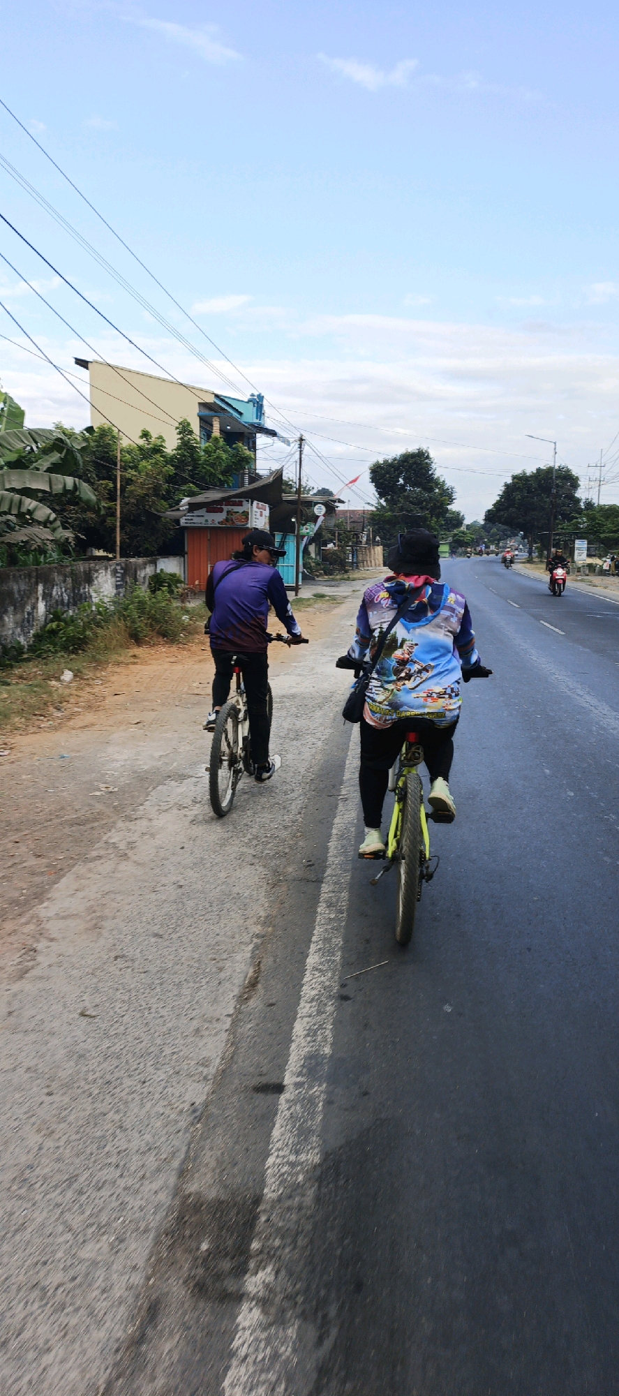 In Kasiyan, Indonesia, two individuals are cycling on a paved road. The person in front is wearing a purple long-sleeved shirt and black pants, with a black cap and sunglasses. They are riding a bicycle with white tires and black rims. To their right and slightly ahead is another person wearing a colorful long-sleeved jacket with a graphic print, black pants, and a black hat. This cyclist is also riding a bicycle, which has yellow rims and black tires. They are both on the right side of the road, with the left side of the road being a dirt path. In the background, there are buildings, trees, utility poles, and other vehicles, including motorcycles, on the road. The sky is visible with scattered clouds.