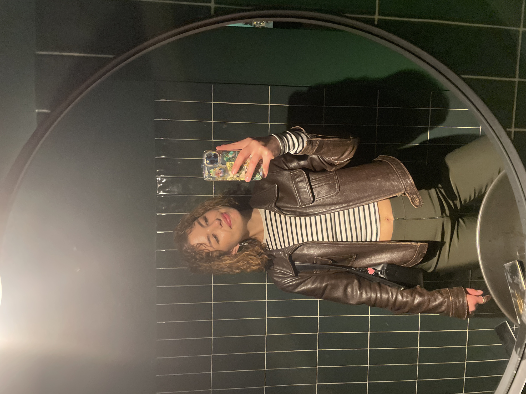 A woman stands in front of a mirror and takes a selfie. The background consists of green-tiled walls. The location could potentially be in Budva, Montenegro. The woman is wearing a leather jacket and a striped shirt. She is holding a smartphone with a patterned case. The oval mirror has a black frame. There is a sink visible in the lower right corner.