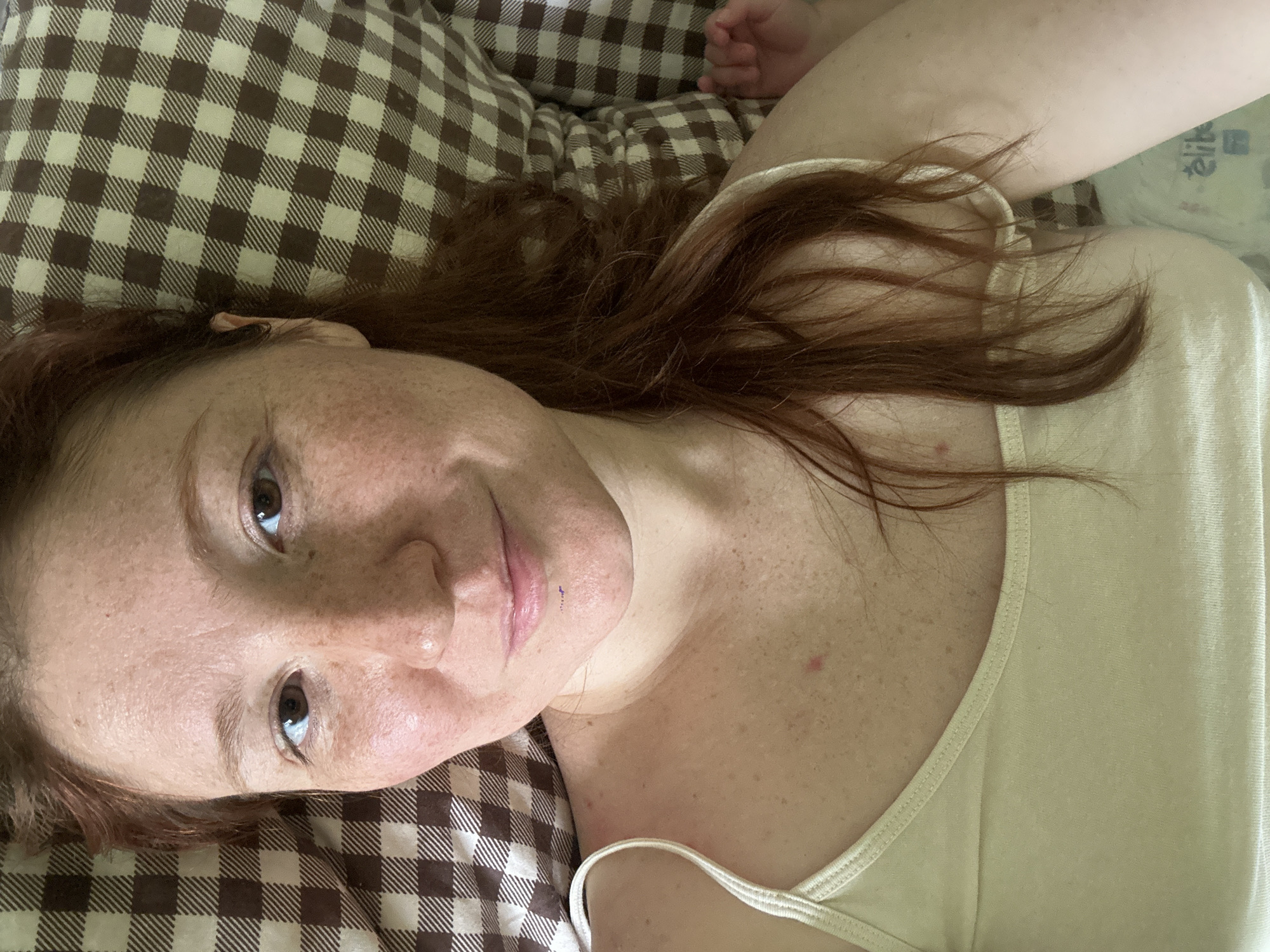 A woman with red hair and freckles is lying down on a brown and beige checkered pillow, wearing a light yellow tank top. She is looking at the camera. Part of a hand is visible in the upper right corner. The background is blurred. There is no indication of the location in the city of Nizhny Novgorod, Russia.