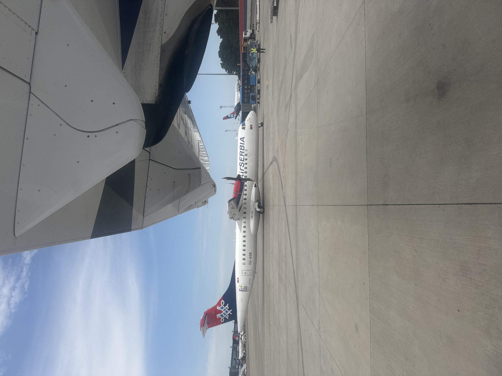 The image captures an outdoor scene at Belgrade Nikola Tesla Airport in Surčin, Serbia. The foreground is dominated by the concrete tarmac and the underside of an airplane's wing. In the mid-ground, an Air Serbia turboprop airplane, registered as YU-ASB, is parked on the tarmac. Its tail features the Air Serbia logo and the "EXPO 2027" emblem. To the right of this aircraft, other aircraft and airport vehicles, including a blue bus and a yellow truck, are visible. A person wearing a high-visibility vest is standing near the bus. The background shows a clear blue sky with scattered clouds and trees lining the airport perimeter. The overall impression is one of an active airport environment under daylight conditions.