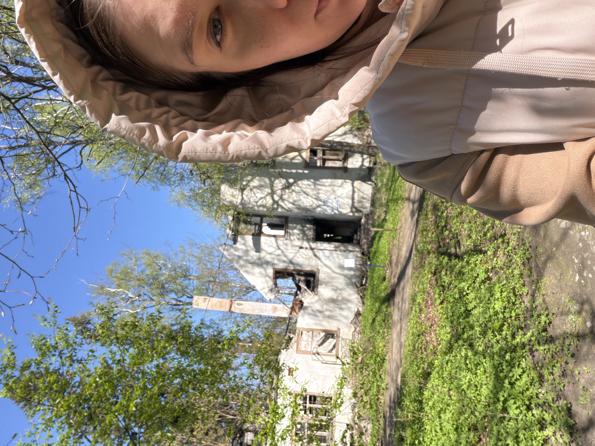 A woman with fair skin and dark hair, wearing a beige hooded jacket, is facing the camera at a tilted angle. She is positioned in front of what appears to be a severely damaged or destroyed building. The building's facade is crumbling, with visible window frames and large, dark openings, likely caused by fire or explosions. A chimney stands next to the building, partially obscured by trees.
The surrounding environment consists of greenery, including grass and trees with fresh leaves, suggesting it is springtime. The sky is clear and blue. The scene suggests a place affected by conflict or disaster. The specific location is Tokkari, Russia.