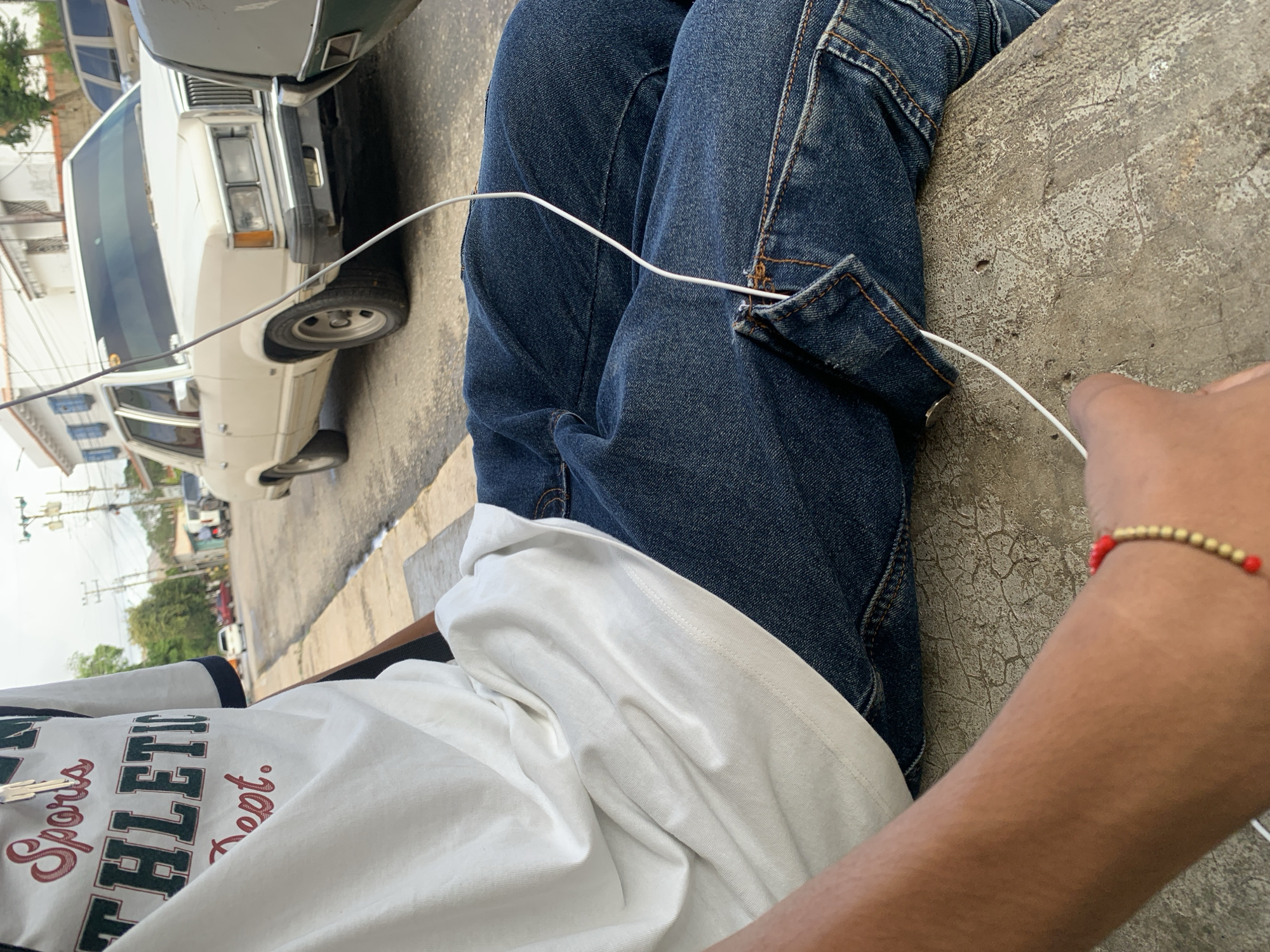 A person is partially visible, seen from the waist down, sitting outdoors. They are wearing dark blue jeans with visible stitching and a white t-shirt. A white cable extends from their lap or pocket area, moving towards their right hand, which is mostly out of frame. On their right wrist, a red and gold beaded bracelet is visible.
In the background, an older model white sedan is parked on the side of a paved street. The car features rectangular headlights and a chrome grille. Other vehicles and buildings are subtly visible further down the street, along with power lines and utility poles, suggesting a typical town environment. The street surface appears to be concrete or asphalt, showing signs of wear and tear.
The lighting indicates a bright daytime setting, possibly sunny. The person's posture suggests a relaxed or resting activity. On the white t-shirt, partial text in red and green lettering reads "Sports" and "ATHLETI C Dept." The overall scene depicts a casual moment on a street in Santa Ana, Venezuela.
