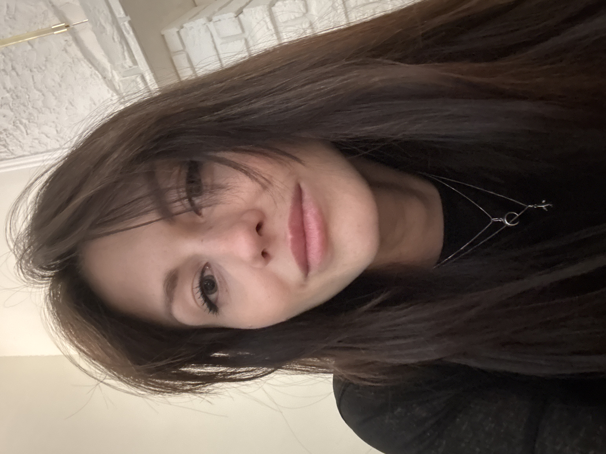 A close-up, rotated portrait shows a young woman with long, dark brown hair that has bangs. Her head is tilted, and her hair falls around her face, slightly obscuring the left side of her forehead and the corner of her left eye. She has light-colored eyes and a gentle, closed-mouth smile. Her lips appear to have a natural pink hue. She is wearing a dark-colored top, possibly black, and a thin, silver-toned necklace with a small, circular clasp or charm visible near her collarbone. The background is out of focus, showing a textured, light-colored surface, possibly a wall or ceiling, with some faint architectural details. The lighting is soft, highlighting her facial features. The overall impression is a casual, intimate portrait. No visible text is present.