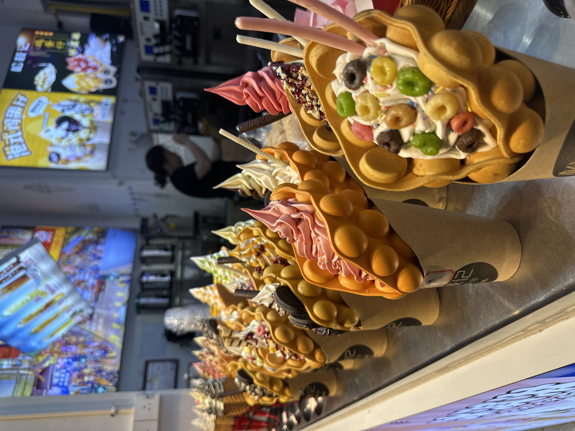 The image depicts a display of bubble waffle cones filled with various ice cream flavors and toppings, such as colorful cereal and chocolate sticks, likely at a street food stall. The bubble waffles are held in paper cones and arranged on a reflective metal surface. A woman in a black shirt can be seen in the background, possibly working at the stall. Two menus are displayed behind the waffle cones. Based on the menus shown, the location is in Furong, China.