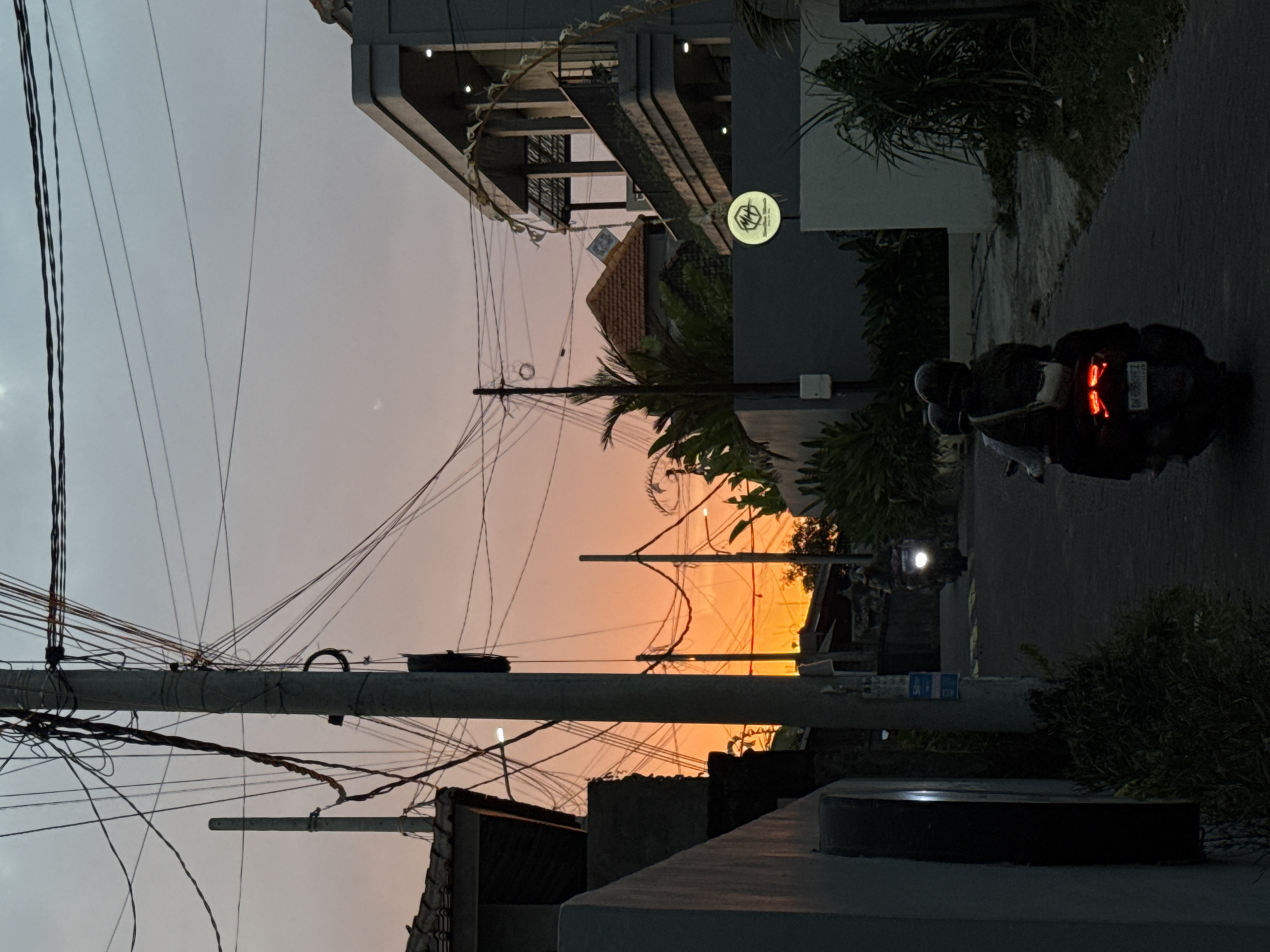 The image depicts a street scene in Kuta Selatan, Indonesia, at dusk. A vibrant orange and pink sunset is visible in the background, casting a warm glow over the scene. The sky is filled with a complex network of electrical wires crisscrossing overhead, suspended from concrete poles. On the right side of the street, a modern building with multiple levels and balconies is prominent, featuring subtle lighting on its exterior. A circular sign with a logo and text is mounted on a wall in front of this building.
The street itself is paved and curves slightly. Two motorcycles are visible on the road. In the foreground, a motorcycle with two occupants, both wearing helmets, travels away from the viewer. The motorcycle's taillights are illuminated, showing a distinct brake light pattern. Further down the street, another motorcycle is approaching, its headlight illuminated.
The left side of the street features darker buildings and vegetation. Lush green plants and trees are present throughout the scene, adding to the tropical ambiance. The overall impression is of a lived-in, active urban environment transitioning into evening.