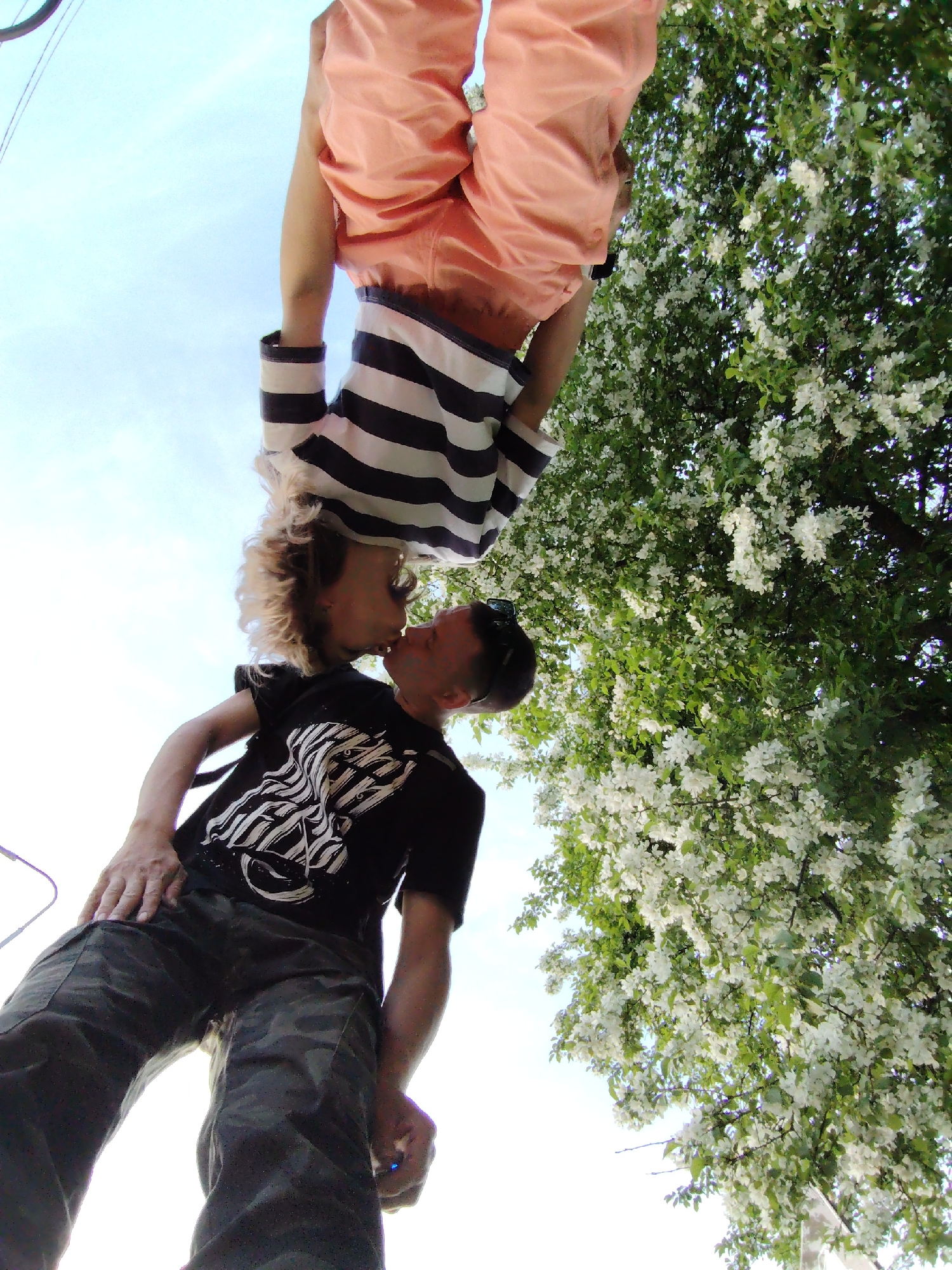 Two people are kissing in an outdoor environment. The person in the foreground is standing and wearing camouflage pants and a black t-shirt with a white graphic. The other person is upside down, wearing a striped shirt and salmon-colored shorts. A tree with white blossoms and a blue sky are in the background. The location is Chaikovsky, Russia.
