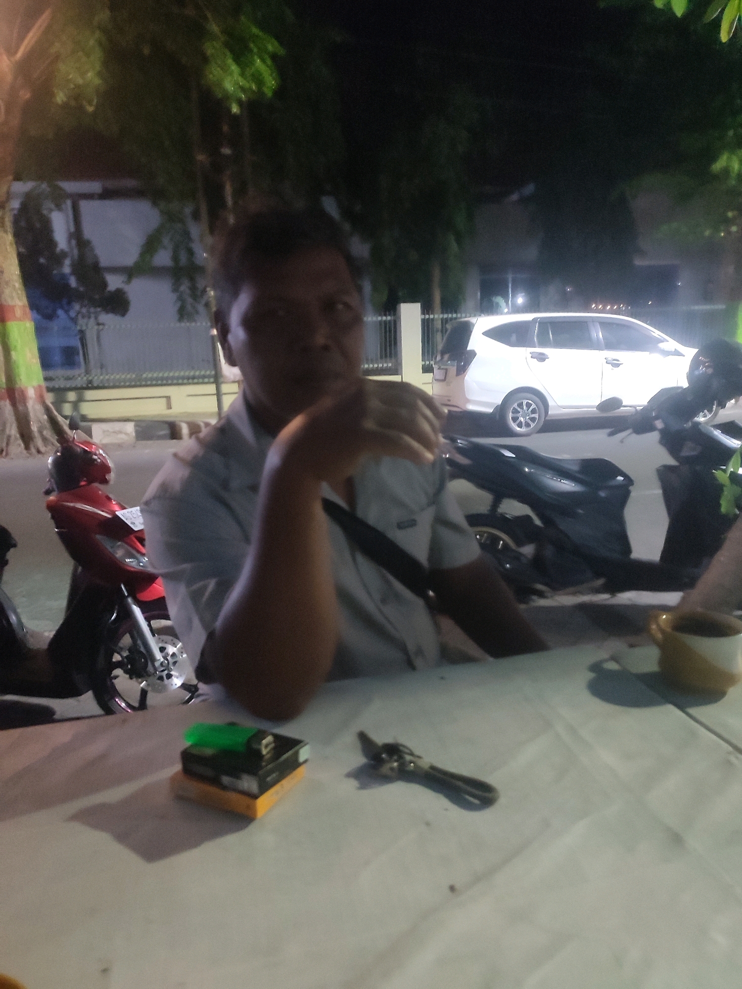A man sits at a table outdoors at night, possibly in Nganjuk, Indonesia, with motorcycles parked behind him and a white car in the background. The man, who appears to be in his late 40s or 50s, wears a light-colored collared shirt. He is captured in a mid-shot, with his right arm resting on the table and his hand near his face. In front of him on the table are a pack of cigarettes and a lighter, and a set of keys. A mug sits further to the right on the table. The lighting suggests it is nighttime, with artificial lights illuminating the scene. The environment includes trees and a building in the background.