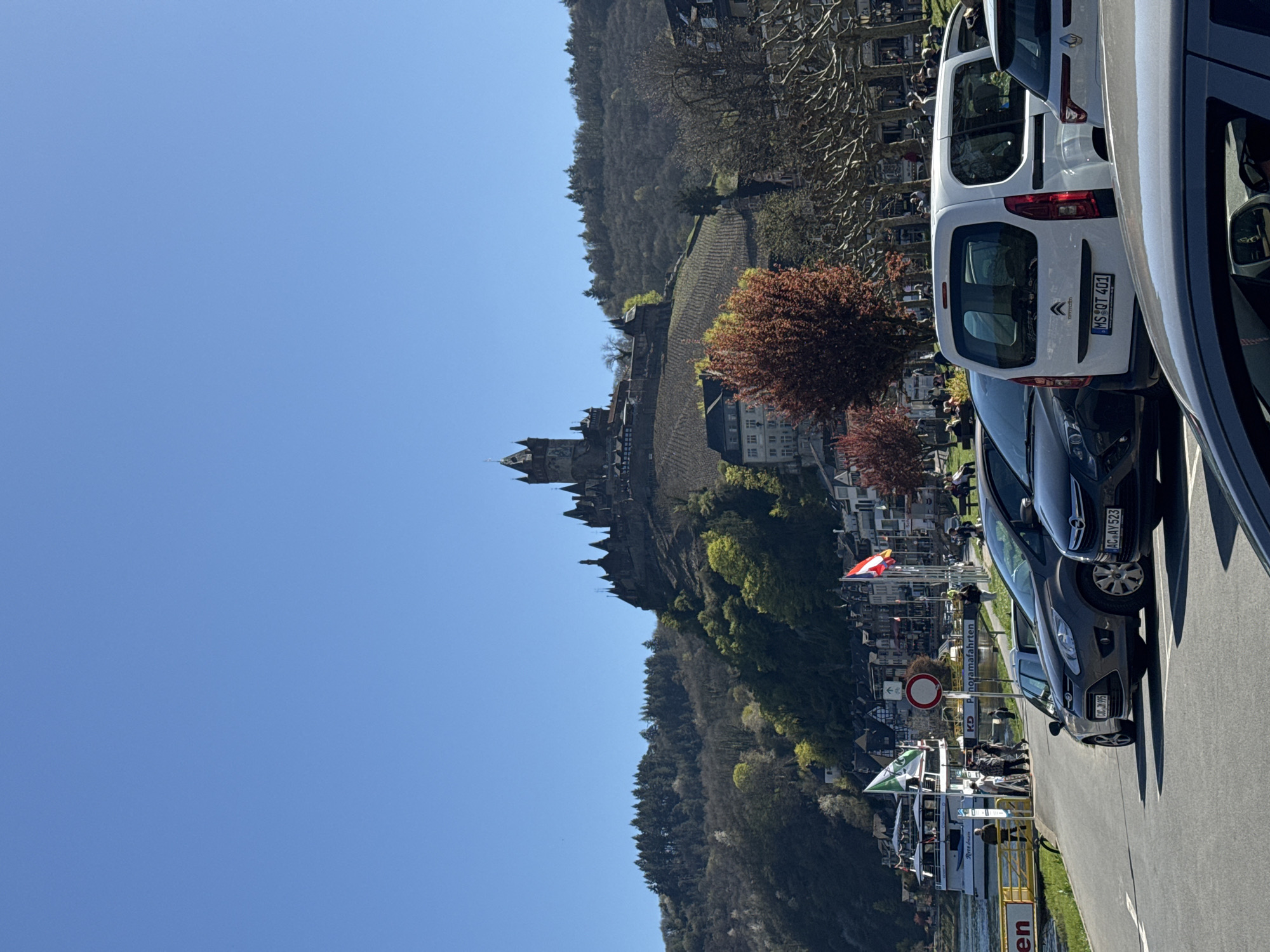 The image shows the town of Cochem, Germany on a sunny day. In the distance, a castle sits atop a hill. The foreground shows a street with parked cars, and a river with a docked tour boat. People are visible near the boat. Flags are flying. A sign with a red circle and horizontal line, indicating a no-entry zone, is visible near the boat. The cars have license plates beginning with "MS" and "AC". A hillside covered with trees is also visible.