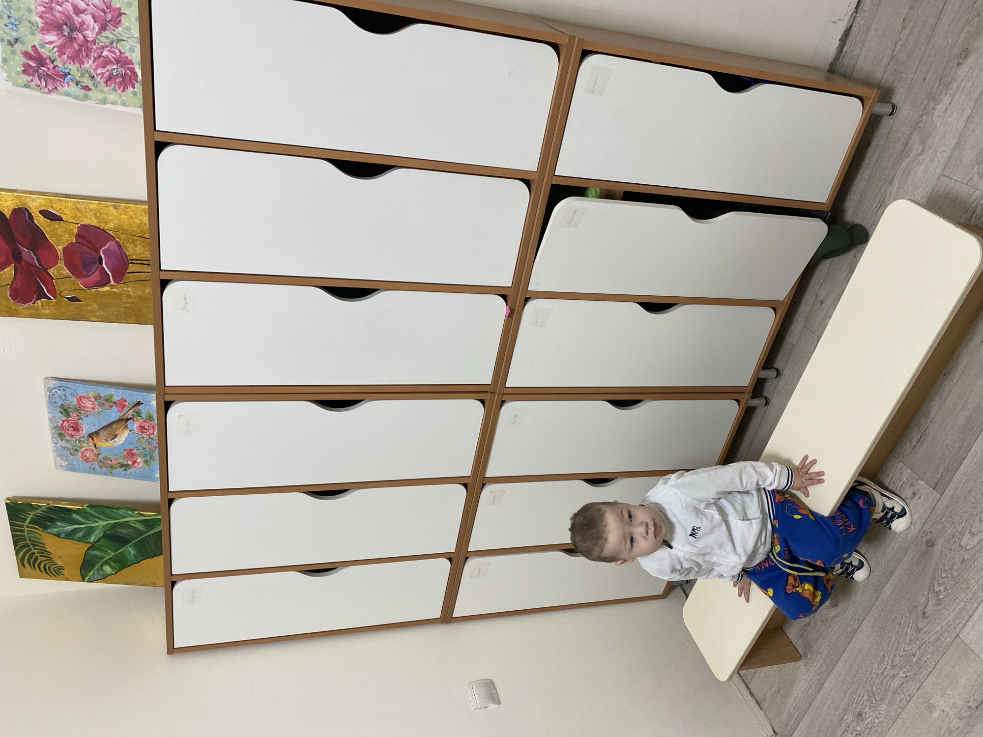 The scene depicts an interior view of a children's changing room or cloakroom within a kindergarten or preschool in the city of Sochi, Russia. A young child, appearing to be a toddler, with short light brown hair, sits centrally on a light-colored wooden bench. The child wears a white zippered track jacket with a "NYC" emblem, blue sweatpants adorned with cartoon characters, and sneakers. The child's gaze is directed slightly to the left of the frame.
Behind the child and extending across the frame is a two-tiered wooden locker unit with white doors, each featuring a curved cutout handle. There are twelve individual compartments in total, six on the top tier and six on the bottom. Several locker doors have names written in Cyrillic on white tape, such as "Мария" (Maria), "Ксения" (Ksenia), "Полина" (Polina), "Леонид" (Leonid), "Егор" (Egor), "Михаил" (Mikhail), and "Богдан" (Bogdan), indicating individual storage spaces for children. One locker door on the bottom tier is slightly ajar, revealing a dark green item inside. The unit rests on short silver-colored cylindrical feet.
Above the locker unit, leaning against a plain light-colored wall, are four distinct rectangular paintings: a gold-toned painting with green tropical leaves, a light blue painting with a bird encircled by pink roses, a gold-toned painting with red poppies, and a light green patterned painting with pink flowers. A white electrical switch or outlet is visible on the left wall. The floor is covered with light gray wood-grain laminate or vinyl flooring.
