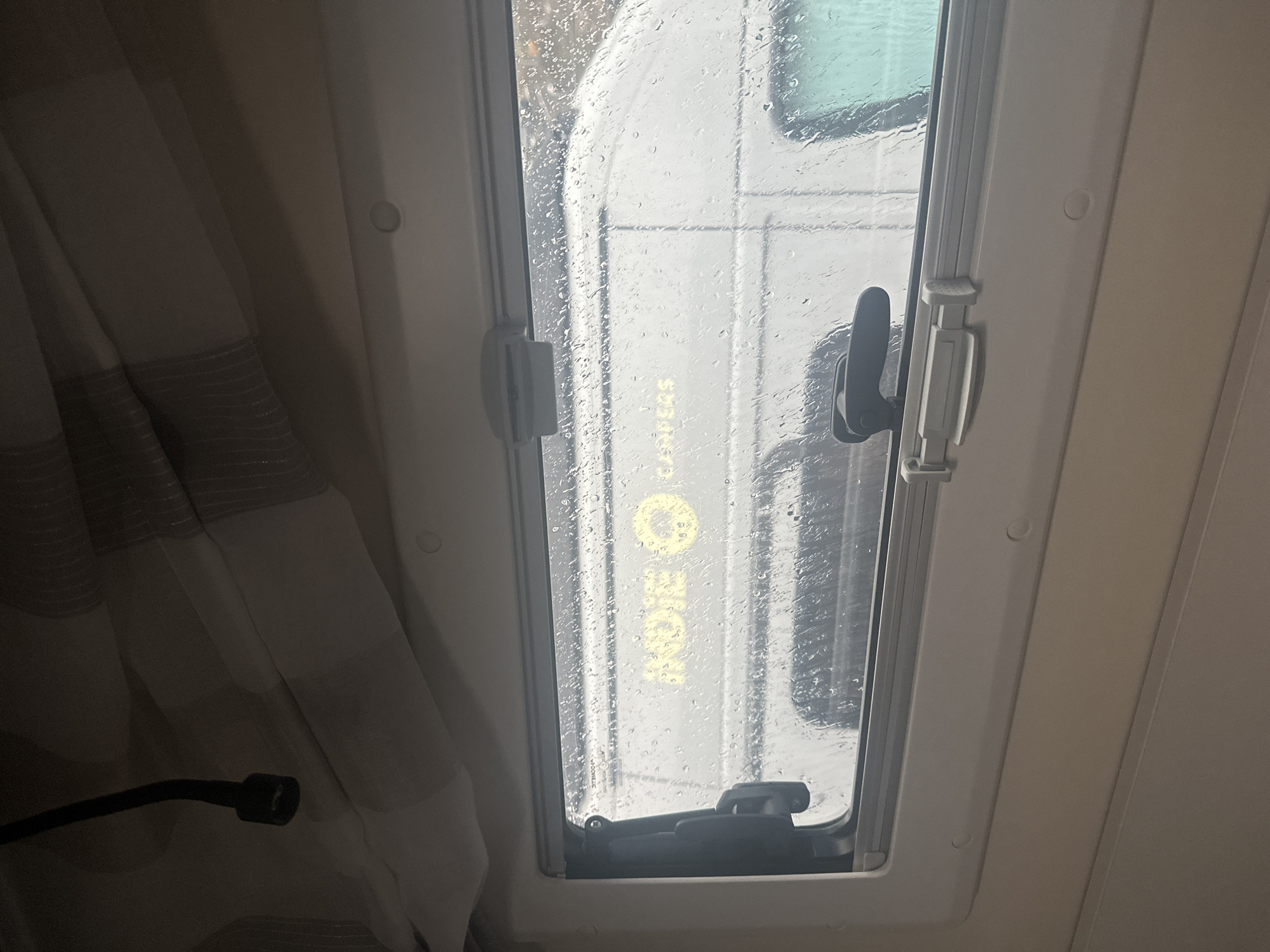 The image shows the interior of a camper van on a rainy day. The view out the window is obscured by raindrops, but the text "ROES CAMPUS" can be seen in yellow on the side of a white vehicle parked outside. A curtain partially covers the left side of the window, and the window's locking mechanism is visible. The location can be inferred to be Montanejos, Spain, based on the text "ROES CAMPUS" seen through the window.