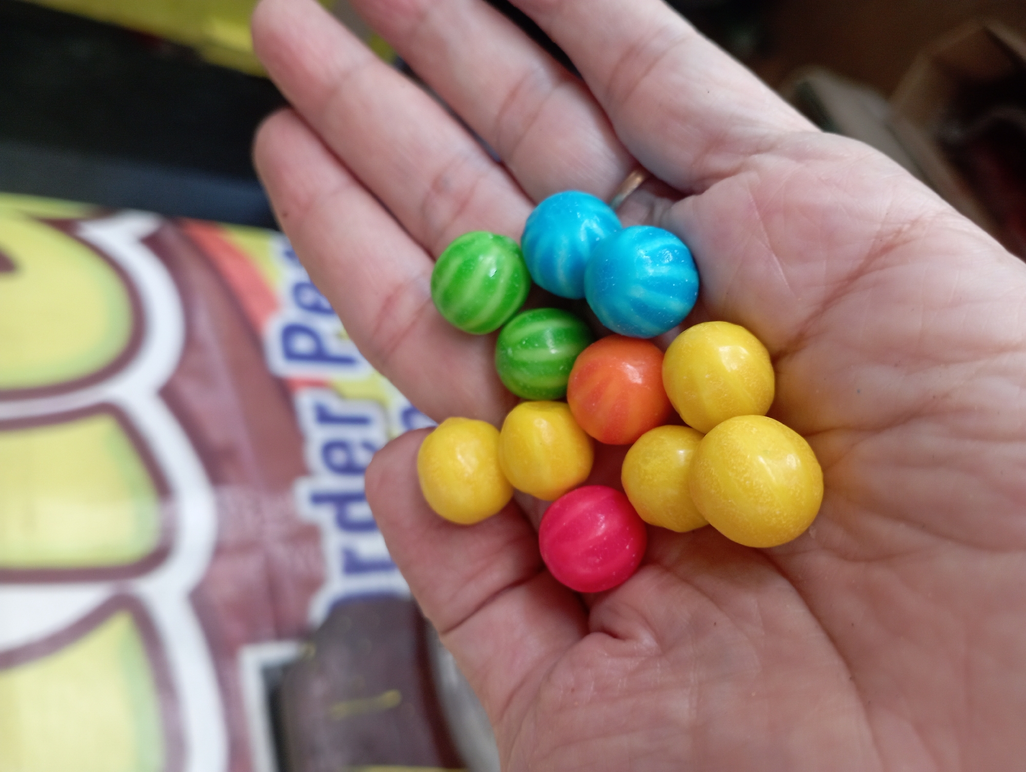 A close-up, vertical shot shows a left hand with a collection of colorful, round candies, possibly gumballs, resting in the palm. The candies are arranged in a cluster, with some overlapping. There are several yellow candies, a pink candy, an orange candy, two blue candies, and two green candies. The skin of the hand is visible, with lines and texture. In the background, out of focus, is a portion of a brightly colored, likely commercial, display. The colors in the background are predominantly yellow and brown, with some blurred text that includes the letters "rder Pe". The lighting appears to be natural, casting a soft glow on the subjects. The scene is devoid of any specific environmental cues that would definitively place it in Sembung Anyar, Indonesia, or any other specific city.