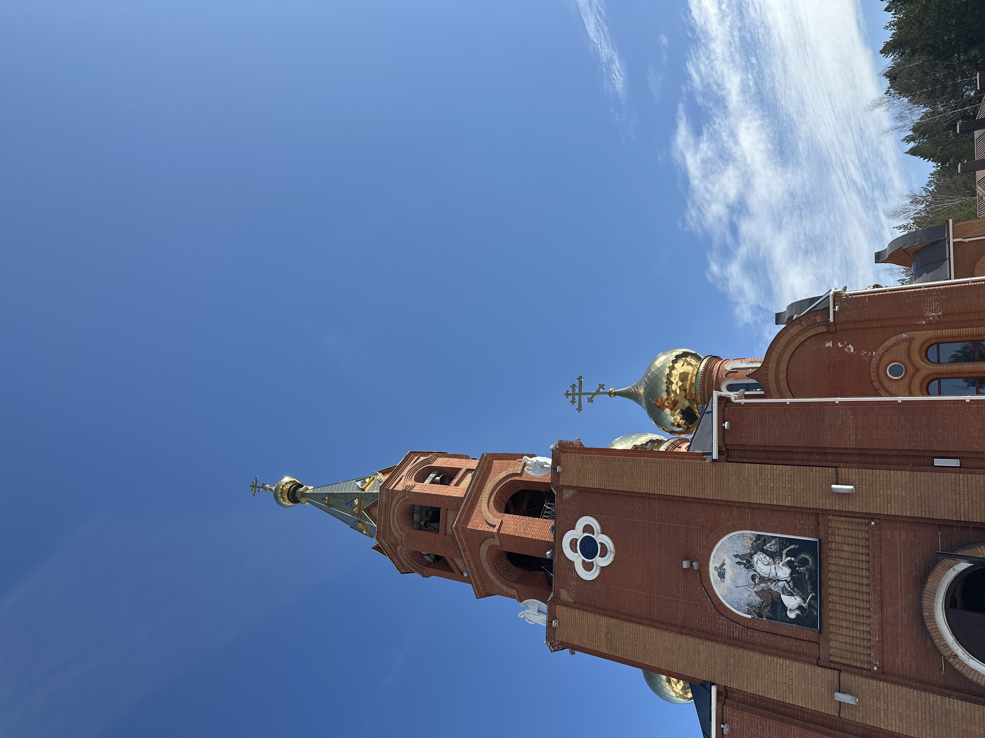 The image presents a low-angle perspective of the Church of the Holy Great Martyr George the Victorious, located at Kamskaya Street 14, Chaikovsky, Perm Krai, Russia. The primary subject is a large Orthodox church constructed from red brick. A tall bell tower rises prominently on the left, featuring multiple arched openings at its upper level, housing bells. Below these arches, two white winged angel statues are positioned on ledges. The bell tower is topped by a slender, conical spire clad in a greenish-gold material, culminating in a golden orb and a Russian Orthodox cross. The main body of the church exhibits multiple golden onion domes, each surmounted by a Russian Orthodox cross. An arched niche on the facade displays a mosaic depicting Saint George on a white horse slaying a dragon. The sky overhead is clear and bright blue, with sparse, wispy white clouds visible along the lower right portion of the frame. A line of evergreen and bare deciduous trees is visible along the lower right horizon, adjacent to the church structure. No individuals or explicit actions are visible within the depicted scene.
