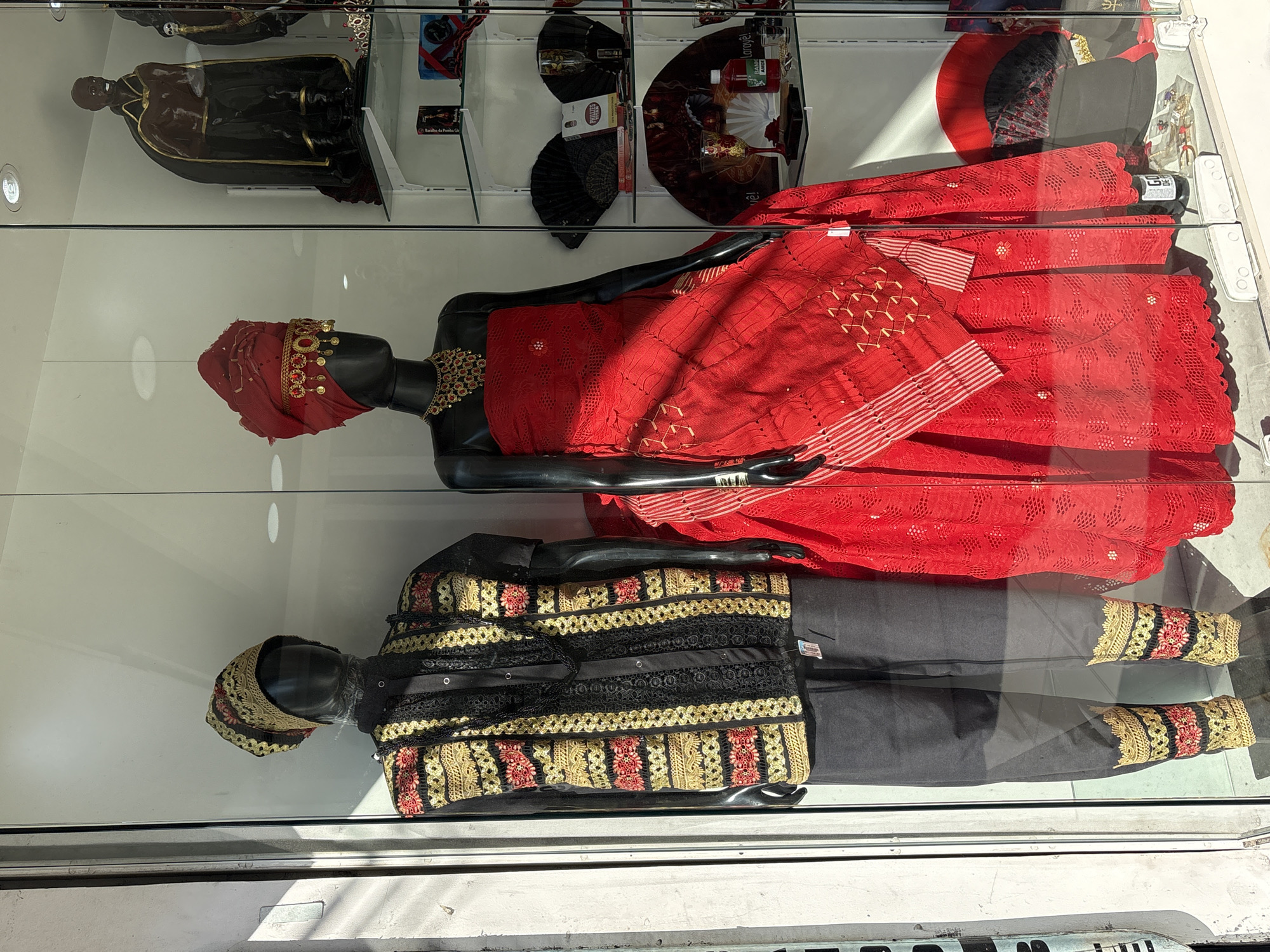 The image shows a display window of a store on Rua 25 de Março in São Paulo, Brazil. The window features two mannequins dressed in traditional clothing. The mannequin on the left wears a black shirt with a patterned vest in gold, red, and black, along with black pants and matching ankle cuffs. It also wears a patterned cap and a beaded necklace. The mannequin on the right is adorned in a vibrant red strapless dress with a matching headpiece and a gold necklace. Behind the mannequins, shelves display various items, including statues, fans, bottles, and decorative objects, suggesting the store sells cultural or religious items. The sunlight casts shadows on the mannequins and the display, indicating it is daytime.