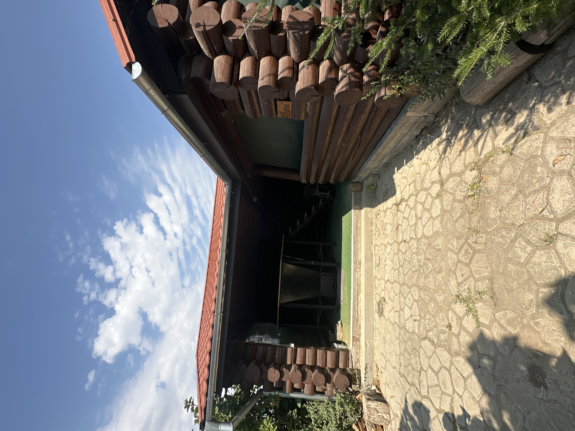 In Marynivka, Ukraine, a log cabin structure with a red tiled roof is seen from a low-angle outdoor perspective on a sunny day. The cabin features a covered open area with a table and what appears to be a metal basin. A sign with Cyrillic script is visible on the side of the cabin. The ground is paved with decorative interlocking stones, and a small green plant is growing through the paving. Shadows of people are cast on the ground, suggesting their presence nearby. The sky above is blue with scattered white clouds.