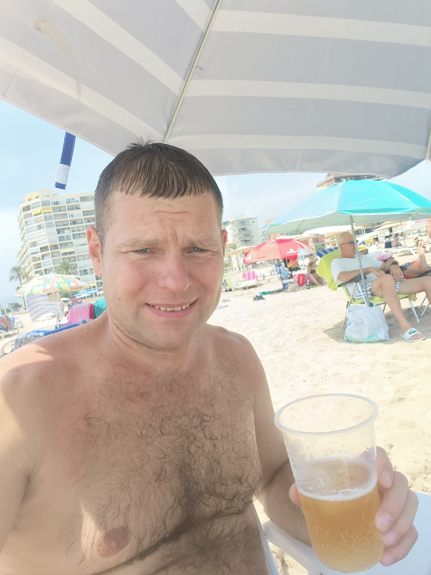 A shirtless man, holding a plastic cup of beer, is taking a selfie on a sunny beach in Gandia, Spain. He is positioned in the foreground, looking at the camera with a smile. Behind him, the sandy beach is populated with beachgoers relaxing under umbrellas, with buildings and palm trees visible in the background. A white and blue striped umbrella shades the man. The overall scene depicts a casual day at the beach.