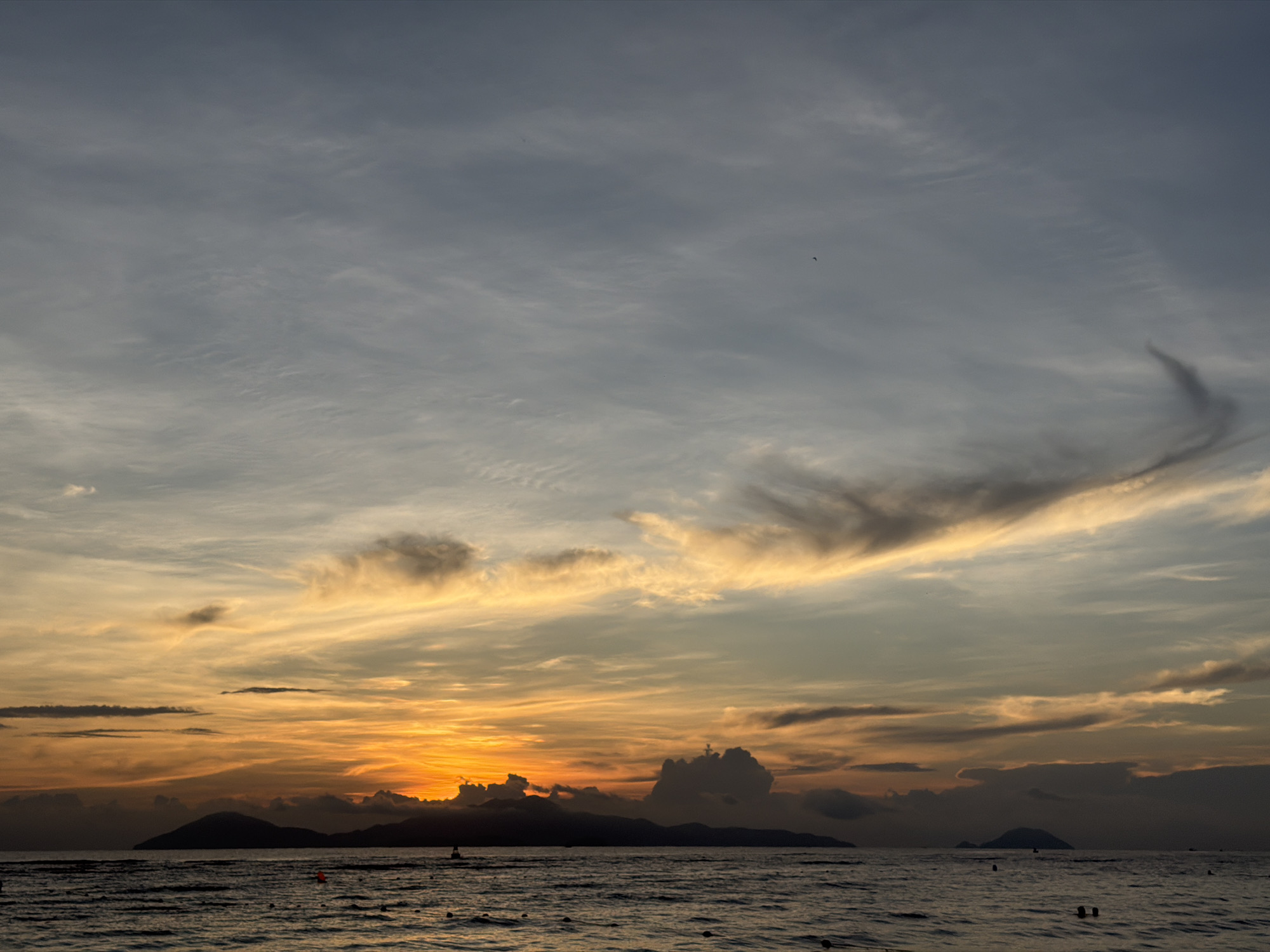 A wide shot captures a serene sunset over the ocean near Hoi An, Vietnam. The sky is a canvas of blues and grays, transitioning to vibrant oranges and yellows near the horizon. Wispy clouds stretch across the sky, catching the fading sunlight. In the foreground, the ocean is dark and choppy, with gentle waves lapping against the shore. Silhouetted against the sunset are several islands and mountains. A few buoys and what appear to be distant swimmers are visible in the water, hinting at a populated beach. The overall atmosphere is tranquil and picturesque.