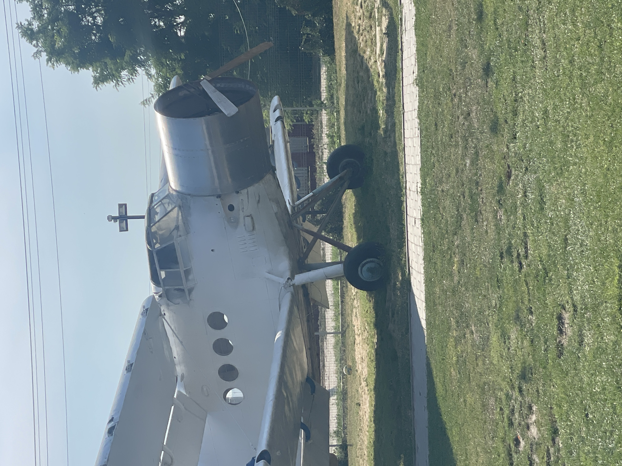 An outdoor, eye-level shot shows a stationary, white Antonov An-2 biplane, facing left. The aircraft is parked on a grassy area adjacent to a paved walkway with a tiled edge. The plane's propeller is visible, as is its engine cowling, cockpit, and part of its wings. A metal fence runs along the background, behind which are trees and a building. A cross-shaped marker is positioned above the cockpit. The sky is clear and blue. The scene is consistent with a static display or a museum exhibit. The location is identified as Zîrnești, Moldova.