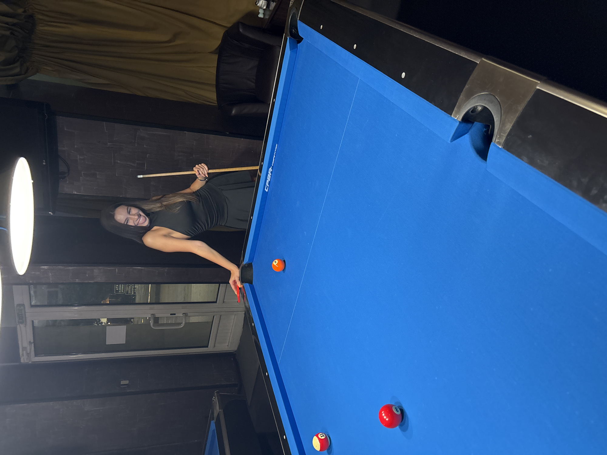 In Vratsa, Bulgaria, a young woman with long dark hair and a black dress is playfully posing by a blue pool table. She is holding a pool cue and sticking her tongue out, her expression suggesting amusement. The room appears to be a dimly lit bar or entertainment venue, with a door visible in the background and a plush armchair. Three billiard balls, a red 3, a red and white 11, and an orange ball, are scattered on the vibrant blue felt of the pool table.