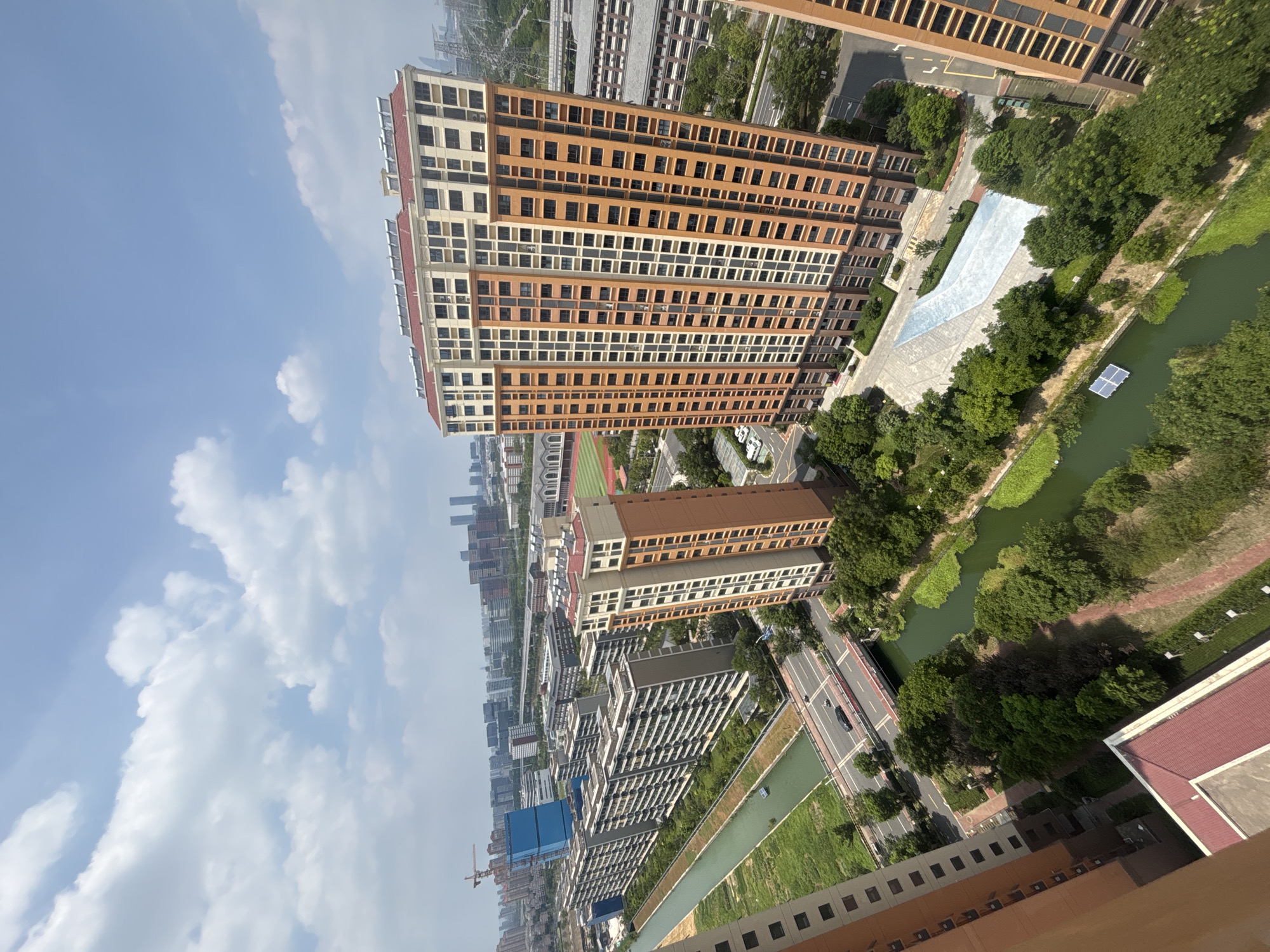 An aerial perspective captures a sunlit urban landscape in Suzhou Industrial Park, China. Dominating the foreground is a tall, modern residential building with a distinctive orange and beige facade. To its left, a series of lower, elongated residential blocks stretches out, bordered by a lush green canal. The canal, reflecting the blue sky and white clouds, winds its way through the scene, with vegetation growing along its banks.
A road with a single black car driving on it runs parallel to the canal, crossed by a red bridge. Beyond the residential buildings, a dense collection of other urban structures is visible, including more apartment blocks, taller office buildings, and a distant view of a stadium with a red track. Construction cranes hint at ongoing development in the area. The sky is a clear blue, punctuated by scattered white cumulus clouds. The overall impression is one of a developed, contemporary urban environment with integrated green spaces.