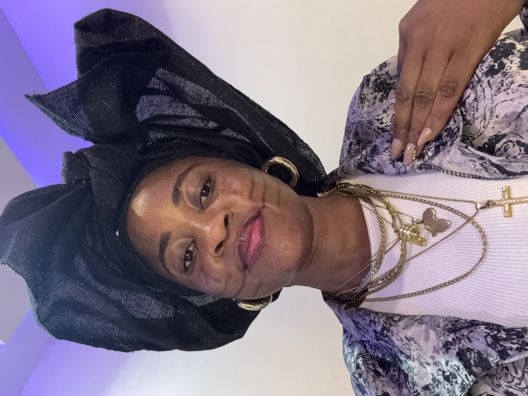 A woman wearing a black headwrap and multiple necklaces is photographed from the chest up in a room with purple lighting. She has subtle face markings and is wearing gold hoop earrings and a printed jacket over a white top. Her right hand is resting on her chest. The image appears to be a selfie taken indoors.