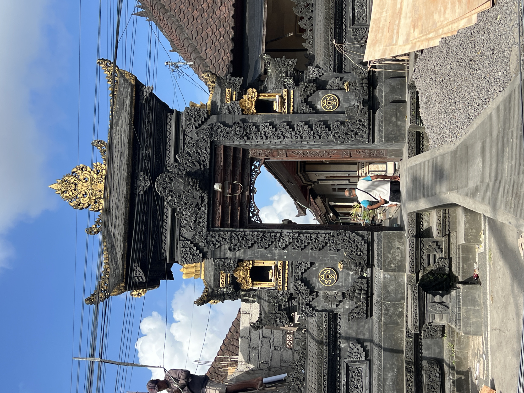 The image shows a highly decorated, black stone Balinese gate or entrance structure with intricate carvings and gold accents, typical of architecture found in the Ubud District, Indonesia. A person wearing a white shirt and blue strap is walking through the gate. To the right, there is an area of gravel. Power lines cross the sky above the structure, and a blue sky with white clouds is visible in the background.