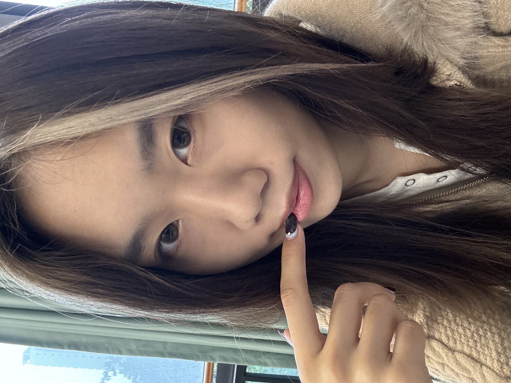 The image displays a close-up frontal portrait of an East Asian female. She possesses long, dark brown hair with a distinct blonde highlight on the viewer's left side, framing her face. Her dark brown eyes are open, directed towards the camera. Her left index finger, adorned with dark nail polish featuring a white crescent near the tip, gently touches the skin below her slightly parted, pink-toned lower lip. She is wearing a light-colored, textured knit garment, likely a sweater or jacket, over a lighter-colored, buttoned undershirt visible at the neckline. A fluffy, light brown collar is visible over her right shoulder (viewer's left). The background indicates an indoor setting within an urban area of Nan'an District, China. A window to the viewer's left provides a partial view of a tall, light-colored building and a dark railing against a light sky, partially obscured by green vertical window coverings.