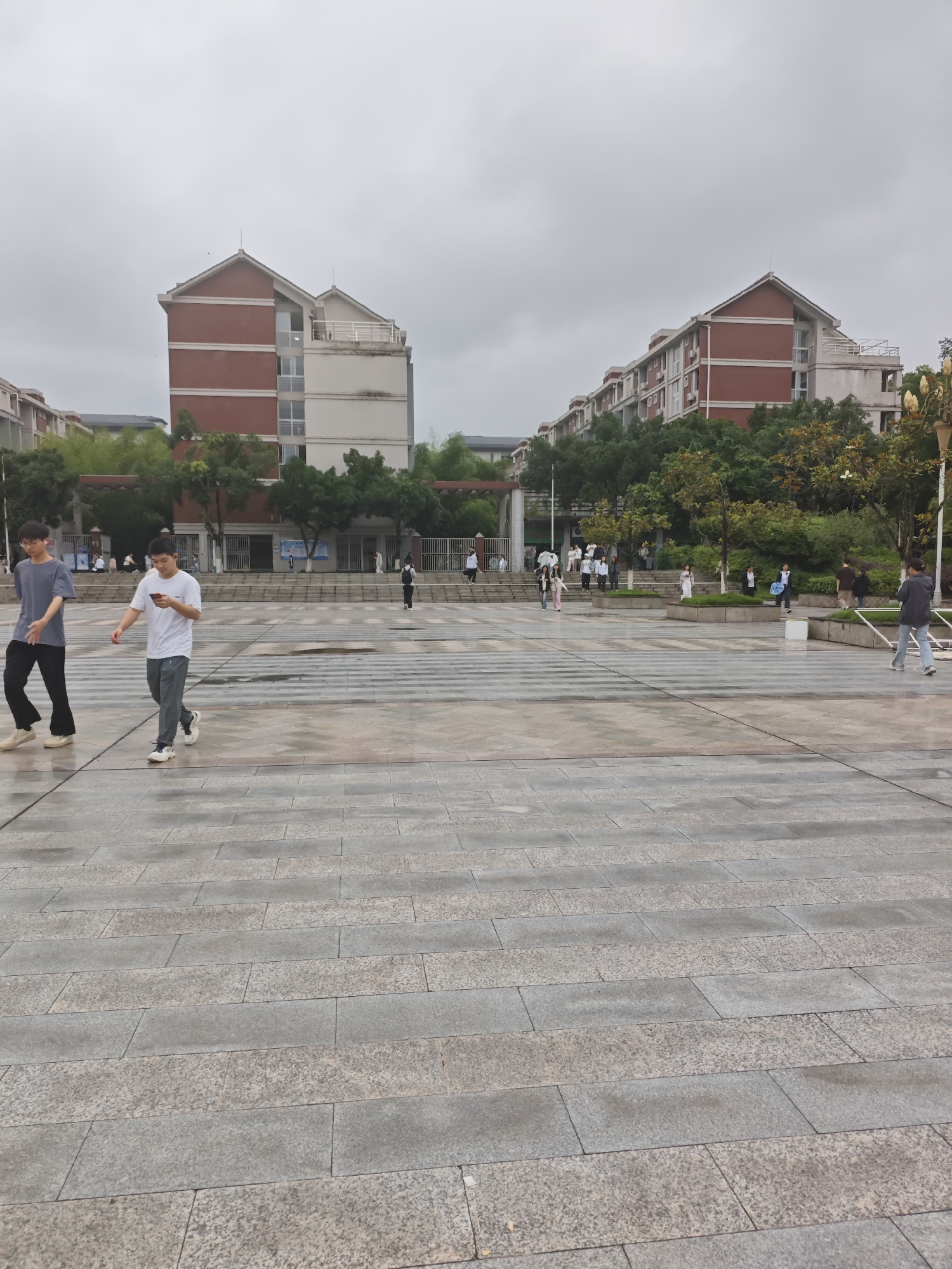 The image depicts an outdoor scene in front of residential buildings under a cloudy sky. In the foreground, a wide paved area with a pattern of large grey stone slabs is visible, with some areas appearing wet. Two young men are walking across this paved area from left to right. The man on the left is wearing a grey t-shirt and dark pants, while the man on the right is wearing a white t-shirt and grey pants and is looking at his phone.
In the middle ground, there are steps leading up to a walkway in front of the buildings. Several people are scattered on these steps and walkways, some are standing, others appear to be walking. One person is holding a white umbrella. The buildings are multi-story structures with red brick accents on the upper floors and white lower floors. They are surrounded by trees and greenery. Further in the background, more buildings and trees are visible, and the sky is overcast. The overall impression is of a university campus or a residential complex on a day with poor weather.