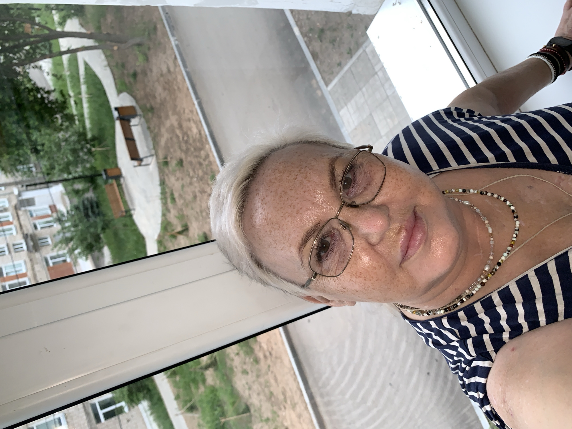 A woman with fair skin, freckles, and short blonde hair is posing for a selfie. She is wearing glasses and a striped blue and white shirt, and several beaded necklaces. The background shows an exterior view through a window, including benches, walkways, trees, and buildings with multiple windows. The presence of buildings of the type shown is common in the city of Chaikovsky, Russia.