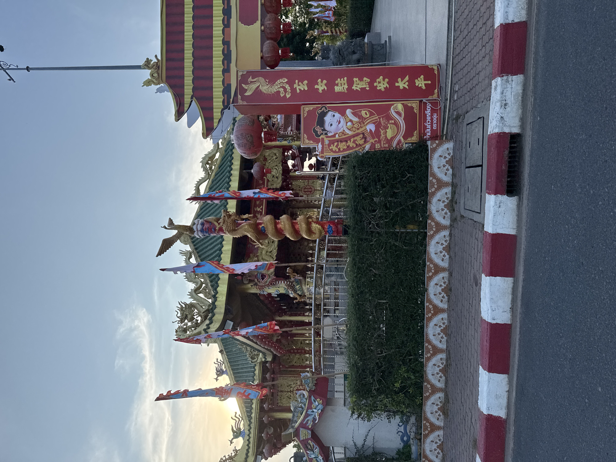 The image depicts a Chinese shrine in Phuket City Municipality, Thailand, with ornate architectural details, including golden dragons and phoenixes. Red lanterns and banners with Chinese characters adorn the shrine. A green hedge runs along the side of the shrine, bordering a street with a red and white curb. A utility pole is visible in the background against a partly cloudy sky.