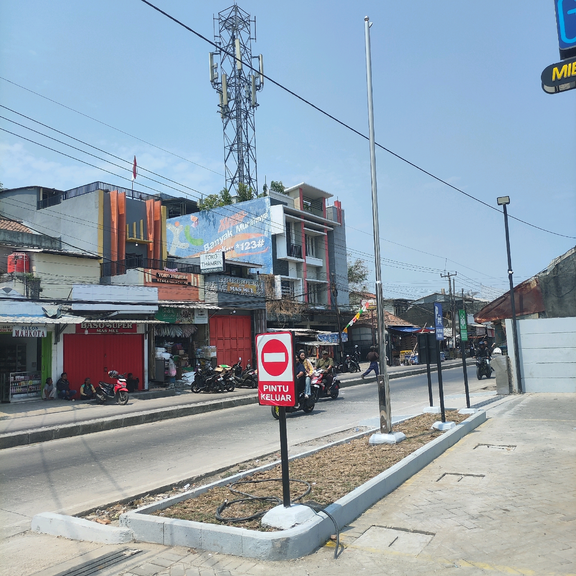 An outdoor, eye-level, daytime shot captures a street scene in an urban environment, likely Southeast Asia, with a clear sky. In the foreground, a grey concrete sidewalk is partially visible, with multiple square metal access covers embedded within. A raised concrete curb separates the sidewalk from a paved road where a red and white sign indicating "PINTU KELUAR" (Exit Door) stands prominently, with a motorcycle carrying two people passing behind it. To the left, a row of shops with red roll-up doors and awnings are situated along the street. Several men are seated outside one of the shops, near a red motorcycle. More motorcycles are parked along the sidewalk. In the background, a large telecommunications tower dominates the skyline, with an Indonesian flag flying atop one of the buildings. Buildings of varying heights and architectural styles line the street, some featuring colorful facades and billboards. Power lines are strung across the scene.