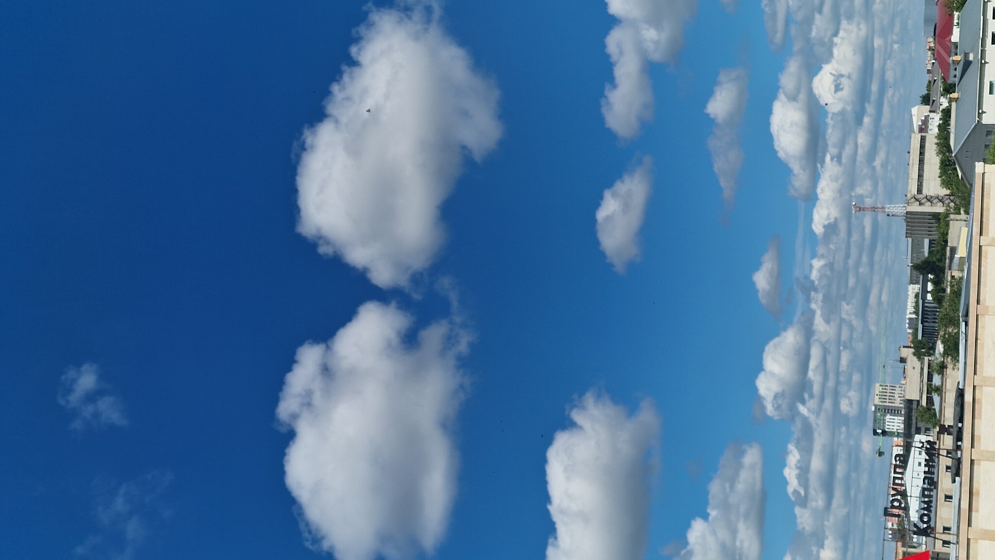 The image shows a bright blue sky with scattered white clouds, typical of a sunny day. In the foreground, a rooftop is visible, with part of a building that has text on it reading "Группа Компаний". Further in the background, a cityscape is depicted with various buildings, including a tall, slender tower with red and white stripes, likely a communication tower. Trees and other structures are also present. The scene suggests an urban environment, possibly within the city of Kostanay, Kazakhstan, given the Cyrillic text visible on the building. There are no people or animals readily discernible in the image.