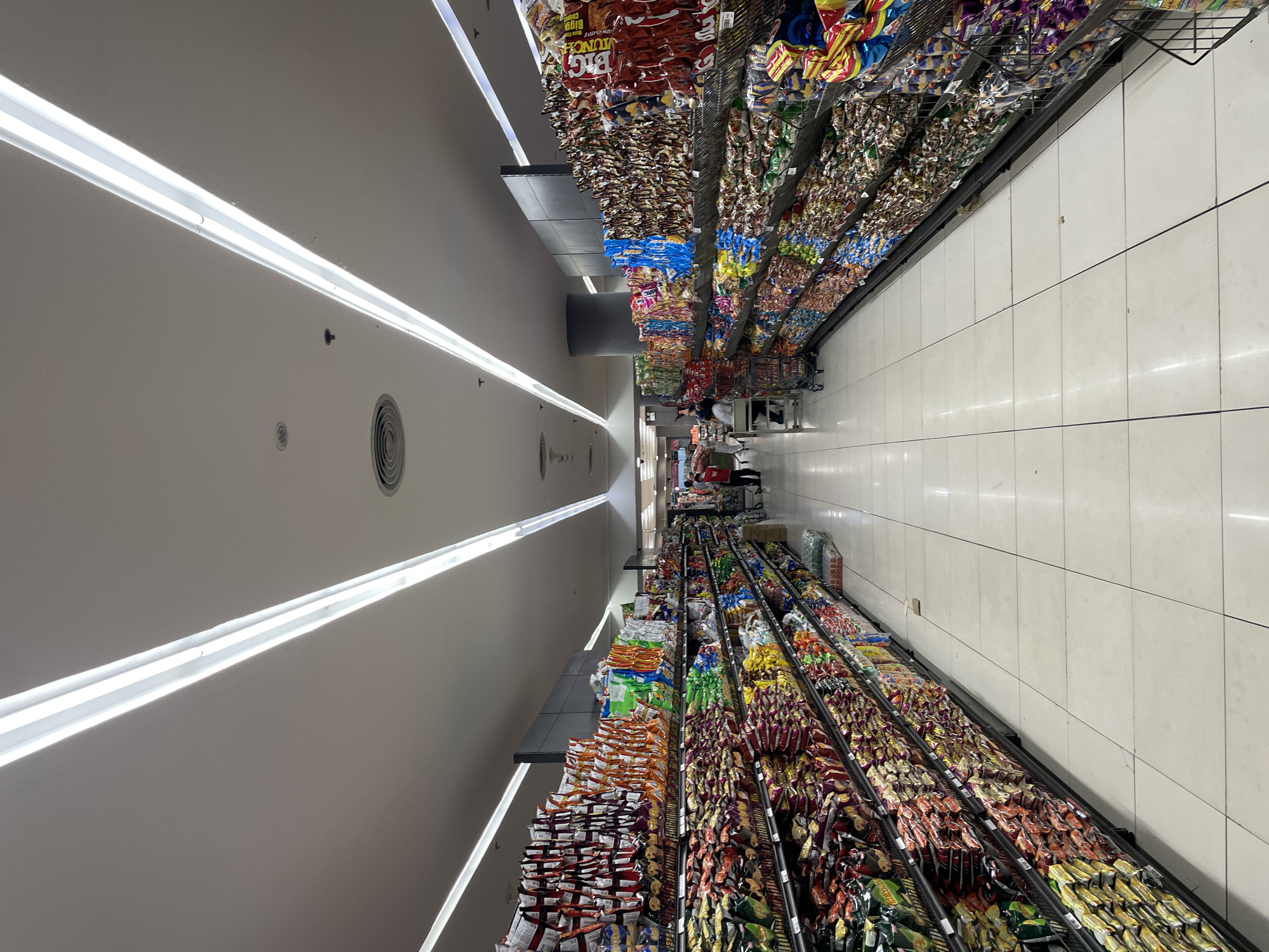 The image shows a wide aisle in a well-lit supermarket. The aisle is lined with tall shelves stocked with a variety of snacks and food items in colorful packaging. The shelves are organized into multiple tiers, with rows of bagged snacks dominating the display. The floor is tiled with large, light-colored squares, creating a clean and expansive look. Two long, illuminated strips run along the ceiling, providing bright, even lighting throughout the space. In the distance, a few shoppers and supermarket staff are visible, with some pushing shopping carts. The overall impression is of a modern, organized retail environment, typical of a supermarket in a developed urban area such as Davao City, Philippines, where such commercial establishments are common.