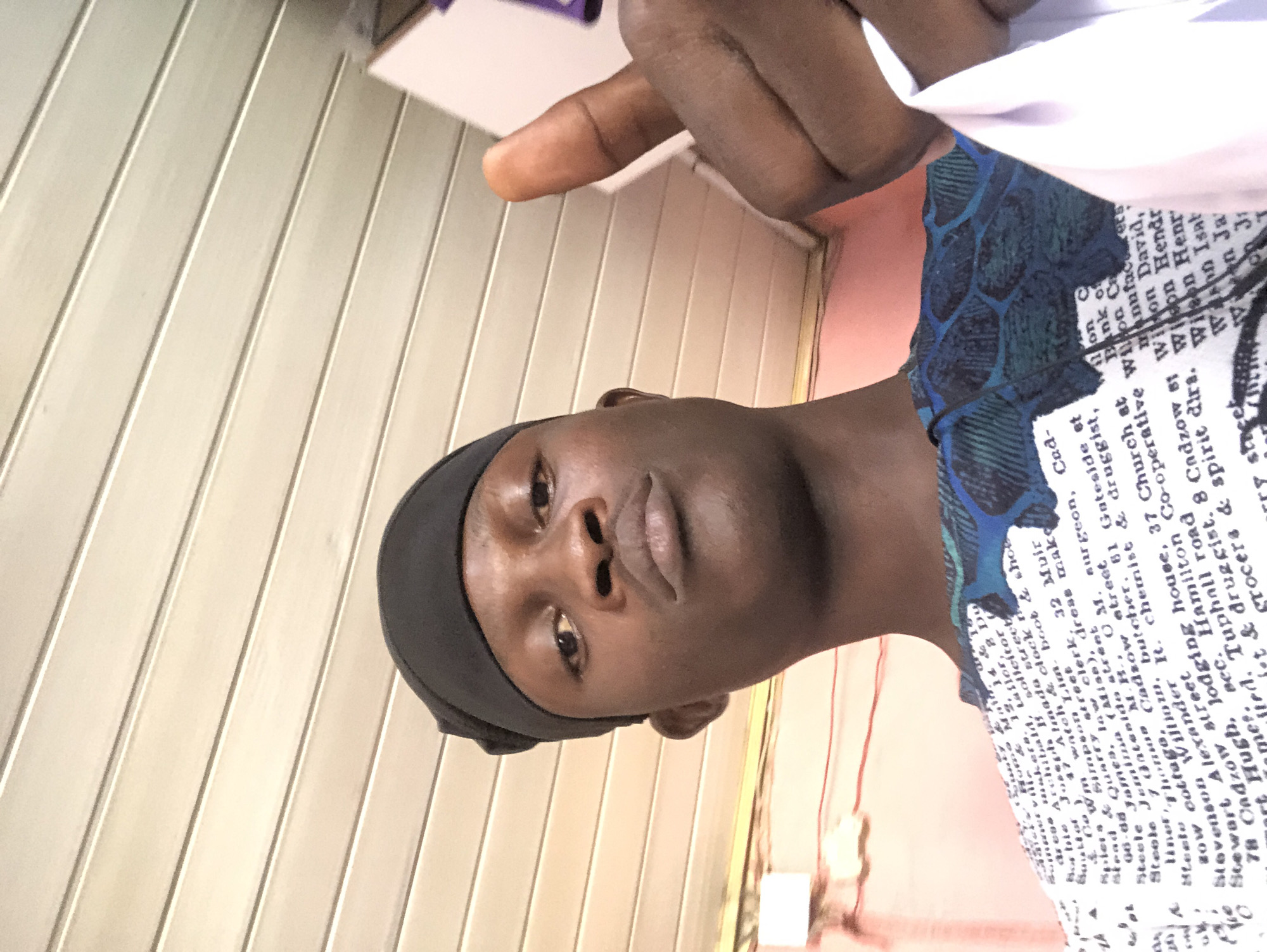 A young Black man wearing a black durag and a patterned t-shirt is captured in a selfie, giving a thumbs-up gesture. The background consists of a light-colored, slatted ceiling and a wall with a peachy-pink hue. Text is visible on the man's t-shirt, appearing to be a list of names and professions, some of which are legible and suggest a directory or business listing. The scene has a casual, indoor setting. The man's skin tone is dark brown, and his facial expression is neutral. The lighting is moderate, casting soft shadows. The image appears to have been taken with a mobile phone.