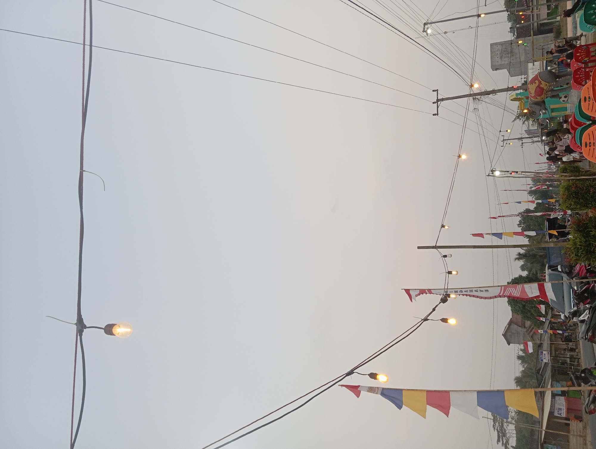 The image depicts an outdoor scene in the city of Way Dente, Indonesia, likely during dusk or early morning given the overcast sky and illuminated string lights. The foreground features several colorful flags, including Indonesian flags, strung across overhead wires. These wires are also adorned with numerous incandescent light bulbs, some of which are lit, casting a warm glow.
In the background, a street is visible with several parked motorcycles and a few vans or cars. Small buildings and structures line the street, suggesting a commercial or residential area. One building has signage that appears to be an ATM. Further in the background, more flags and string lights are visible, extending into the distance.
People are present in the scene, though most are small figures in the background, suggesting a public gathering or marketplace. One group of individuals is gathered near a set of colorful chairs, and a large elephant statue wearing a decorative blanket is visible in the mid-ground. The overall atmosphere appears festive and active, with the lighting and decorations creating a lively ambiance. The environment is characterized by overhead electrical wires, poles, trees, and a generally flat terrain.