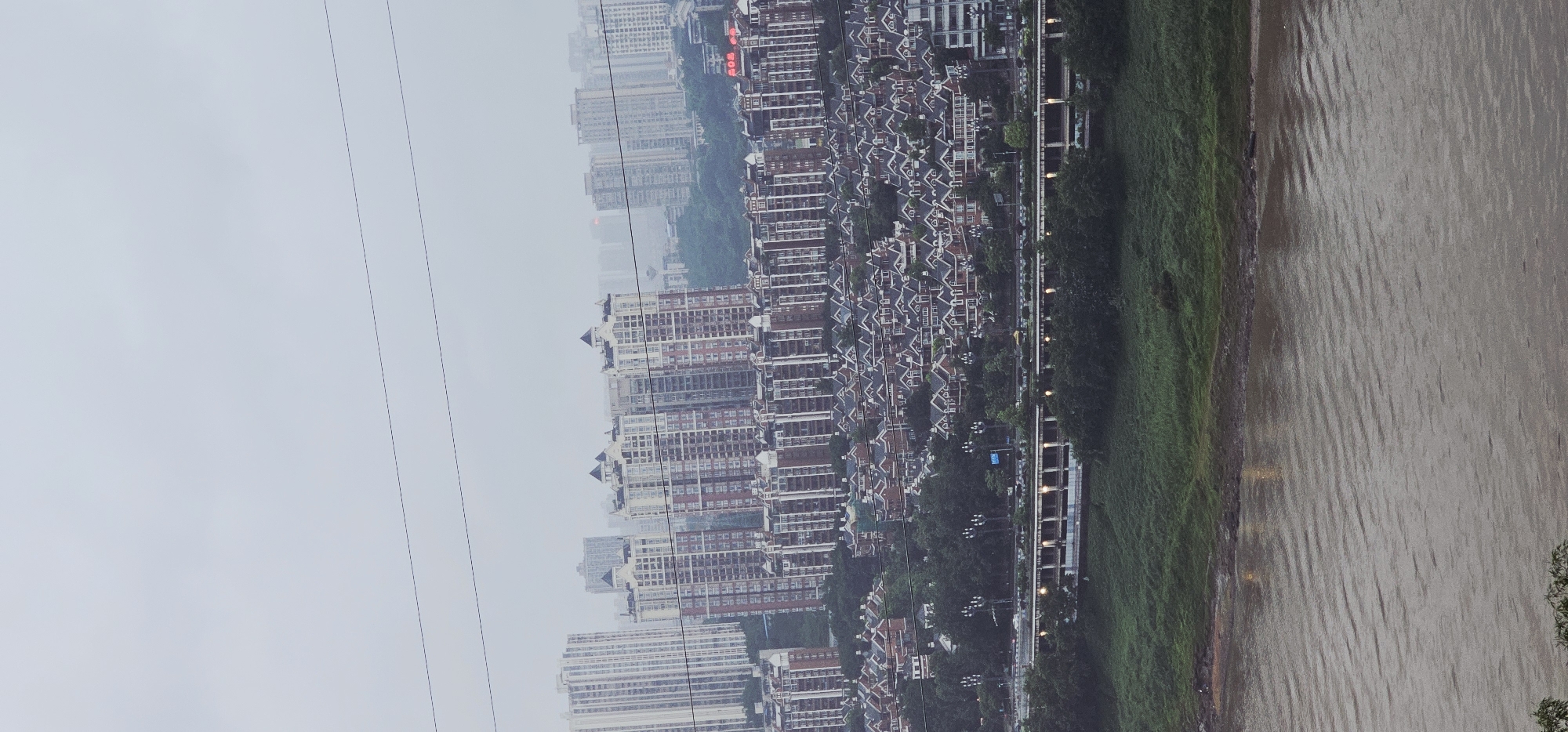 The image displays an elevated, wide-angle view of a dense urban landscape bordering a wide river, characteristic of Yuzhong District, China, under an overcast sky. The top quarter of the frame is dominated by a uniform, light grey sky, horizontally bisected by three dark, parallel overhead lines. Below this, a multi-layered cityscape unfolds. In the mid-ground, numerous tall residential high-rise buildings feature light-colored facades with darker vertical and horizontal accents; several structures are topped with distinctive dark, pointed roof elements. Beneath these, a dense cluster of smaller, multi-story residential buildings with dark sloped roofs and lighter exteriors is arranged on sloped terrain, interspersed with dark green trees. A multi-lane elevated roadway with visible streetlights, some illuminated, traverses horizontally across the lower mid-ground. Parallel and closer to the river is a secondary elevated structure. The bottom third of the image reveals a wide, brownish river with a pronounced rippled surface, separated from the infrastructure by a narrow strip of green, grassy bank. No individuals or specific actions are discernible within the frame.