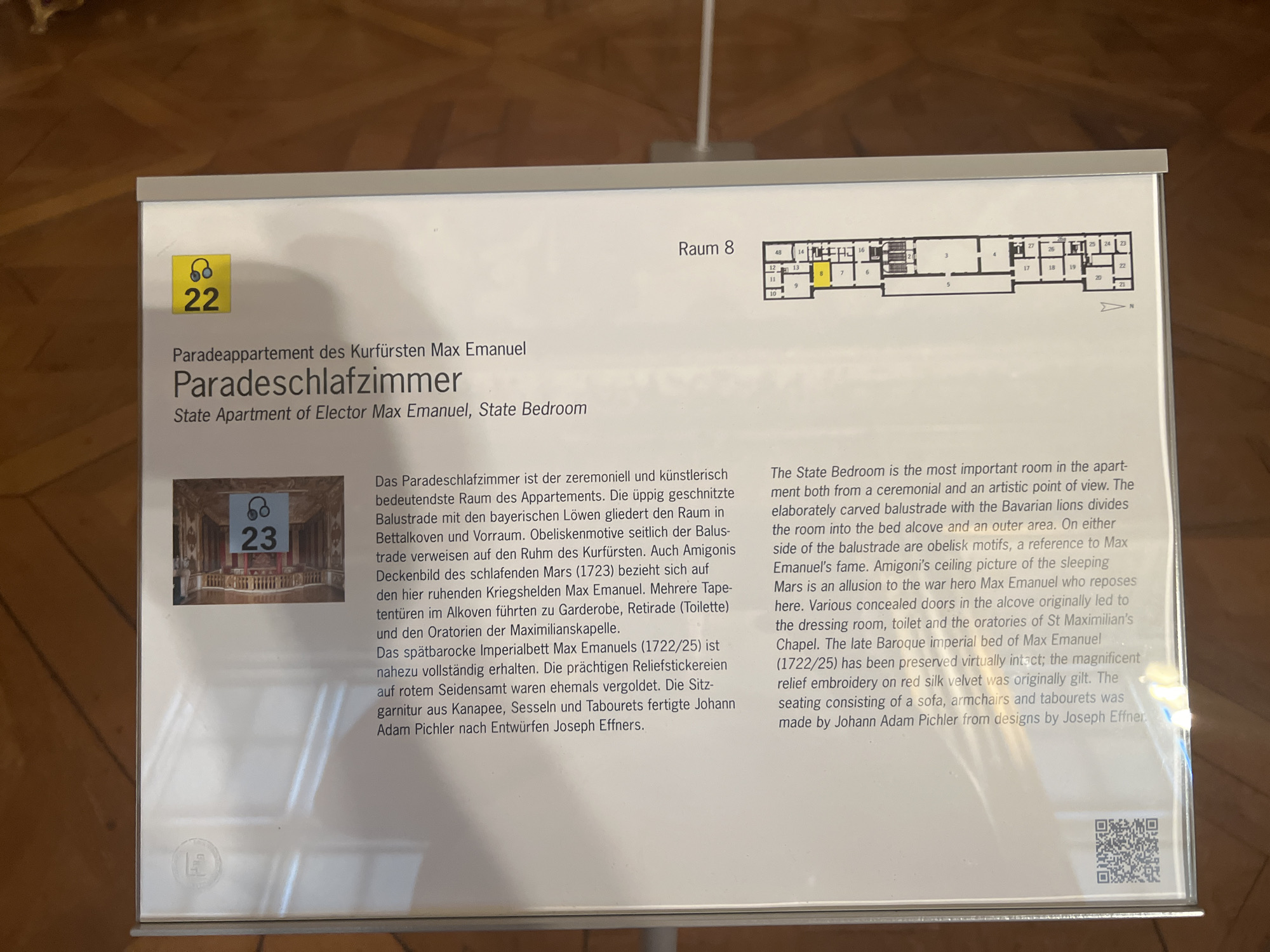 The image shows an informational display board outside a room in the city of Oberschleißheim, Germany. The board has text in both German and English, as well as a floor plan of the area. A photograph of a richly decorated room is also visible on the board, with the number 23 superimposed. The room is identified as the "Paradeschlafzimmer" or "State Bedroom" of Elector Max Emanuel. The floor plan indicates that this room is part of a larger suite, labeled "Raum 8". The context suggests this is a museum exhibit within a palace or historical building.