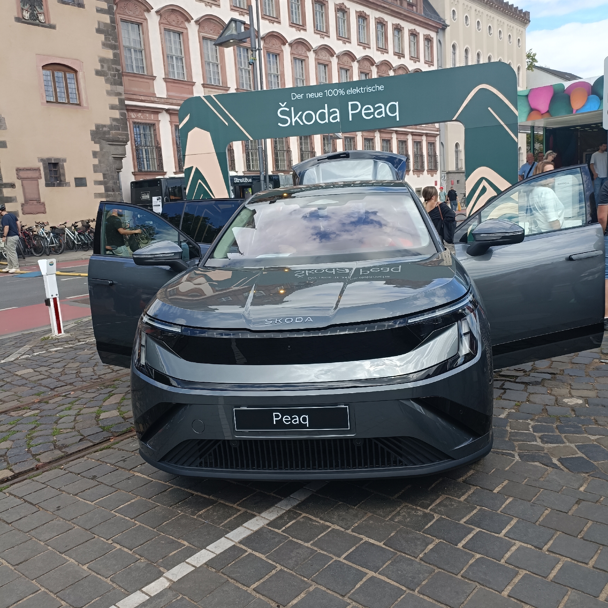 In Frankfurt, Germany, in front of a building with ornate architecture, a gray Skoda Enyaq electric car is parked on a cobblestone street. The car has its doors open, and people are visible inside and around it. A banner overhead reads "Der neue 100% elektrische Skoda Peaq." In the background, a bus is parked, and several bicycles are lined up. The overall scene suggests an outdoor event or exhibition.