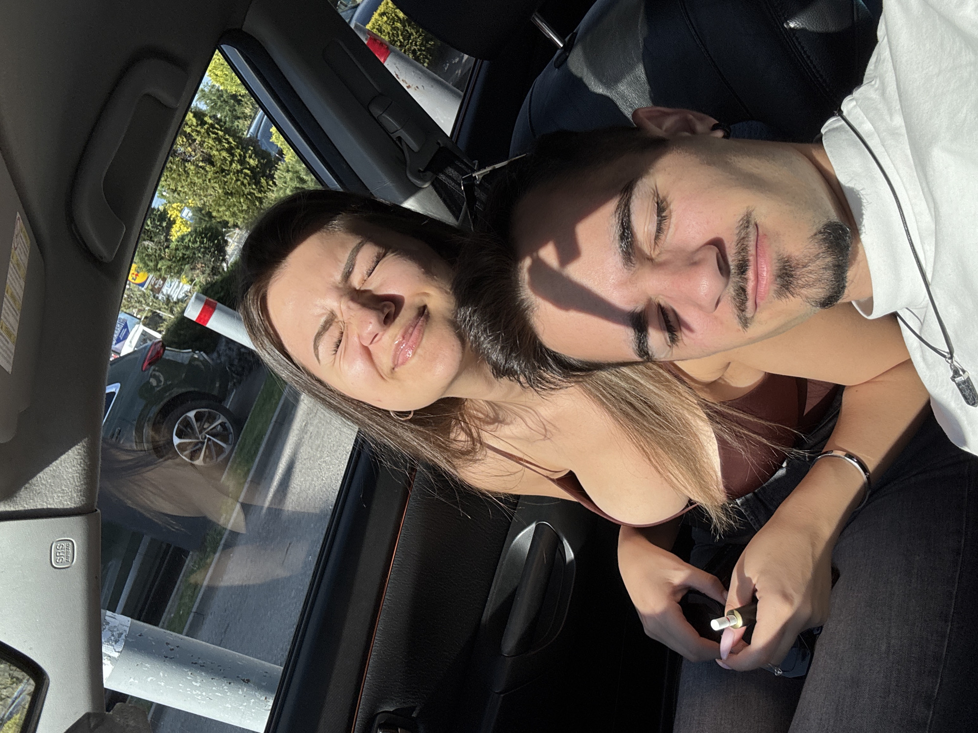 Inside a car, a young woman with long brown hair, wearing a brown top and silver hoop earrings, sits with her eyes closed, her face scrunched slightly in what might be discomfort or contentment. Beside her, a young man with a short beard and wearing a white t-shirt has his eyes closed. The woman is holding a vaping device. The environment outside the car window suggests an urban setting, specifically featuring a Lidl supermarket sign, which might indicate the city of Sofia, Bulgaria.