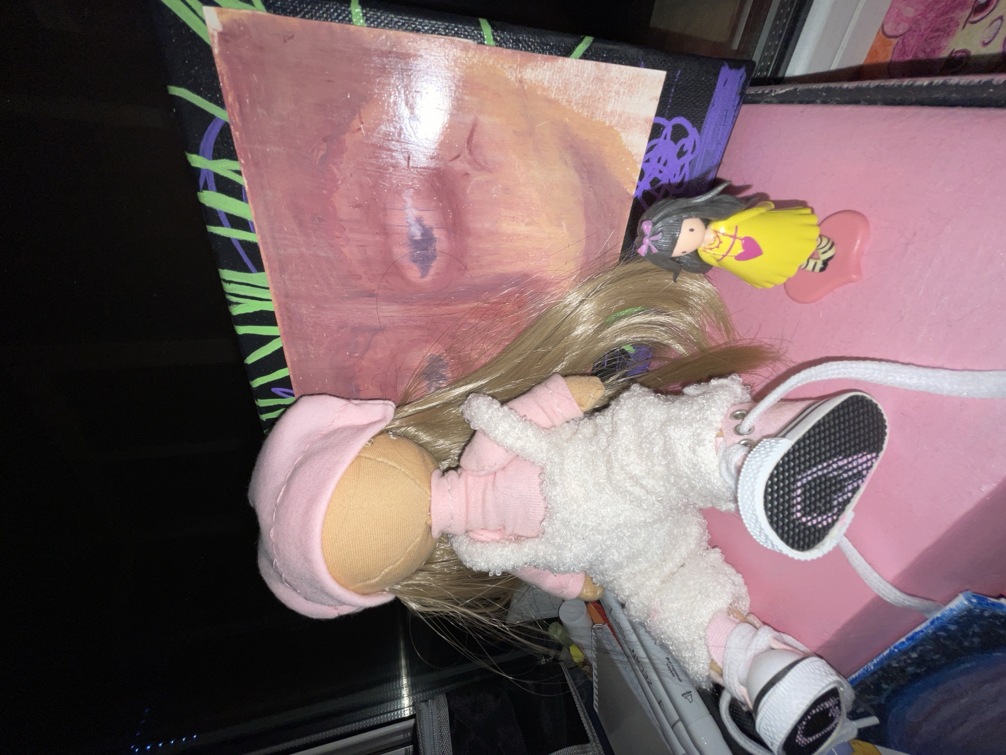 The image shows a large doll wearing a pink beret, pink turtleneck, and white fluffy overalls, with white sneakers. The doll is sitting on a pink surface next to a painting of an abstract face with red and pink tones. A small figurine of a girl in a yellow dress with a pink heart necklace stands to the right of the painting. The background is dark, suggesting it might be nighttime or a dimly lit room. There are some art supplies, including markers, visible to the left of the doll. The overall scene appears to be a setup for a creative project or display. The image does not contain any elements that indicate a specific location in Moscow, Russia.