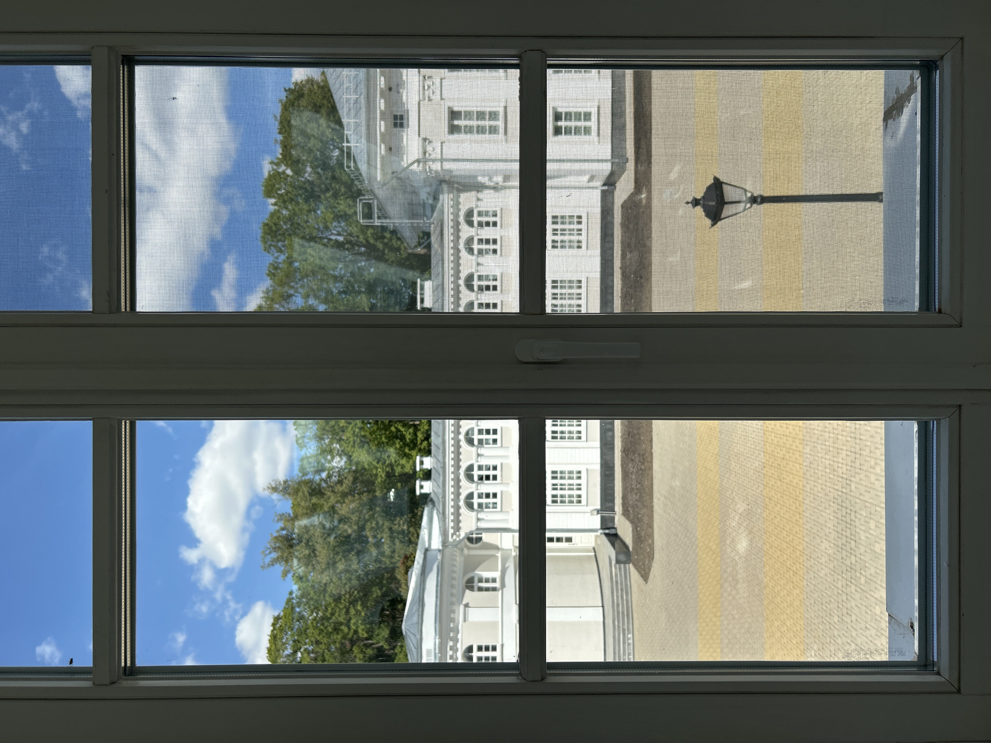 This image, taken from inside a building looking through a window, shows a view of a courtyard with a white, classical-style building in the background. The building has multiple arched windows and decorative architectural elements. Trees are visible behind the building, and the sky above is blue with scattered white clouds. The courtyard pavement is a mix of yellow and grey rectangular bricks. A black lamppost stands in the right-hand section of the courtyard. The window itself is divided into two vertical panes, with a screen visible on the right pane. The scene appears to be set in a park or the grounds of a historical estate. The specific location in Добасненскі сельскі Савет, Belarus, is not discernible from the visual information provided.