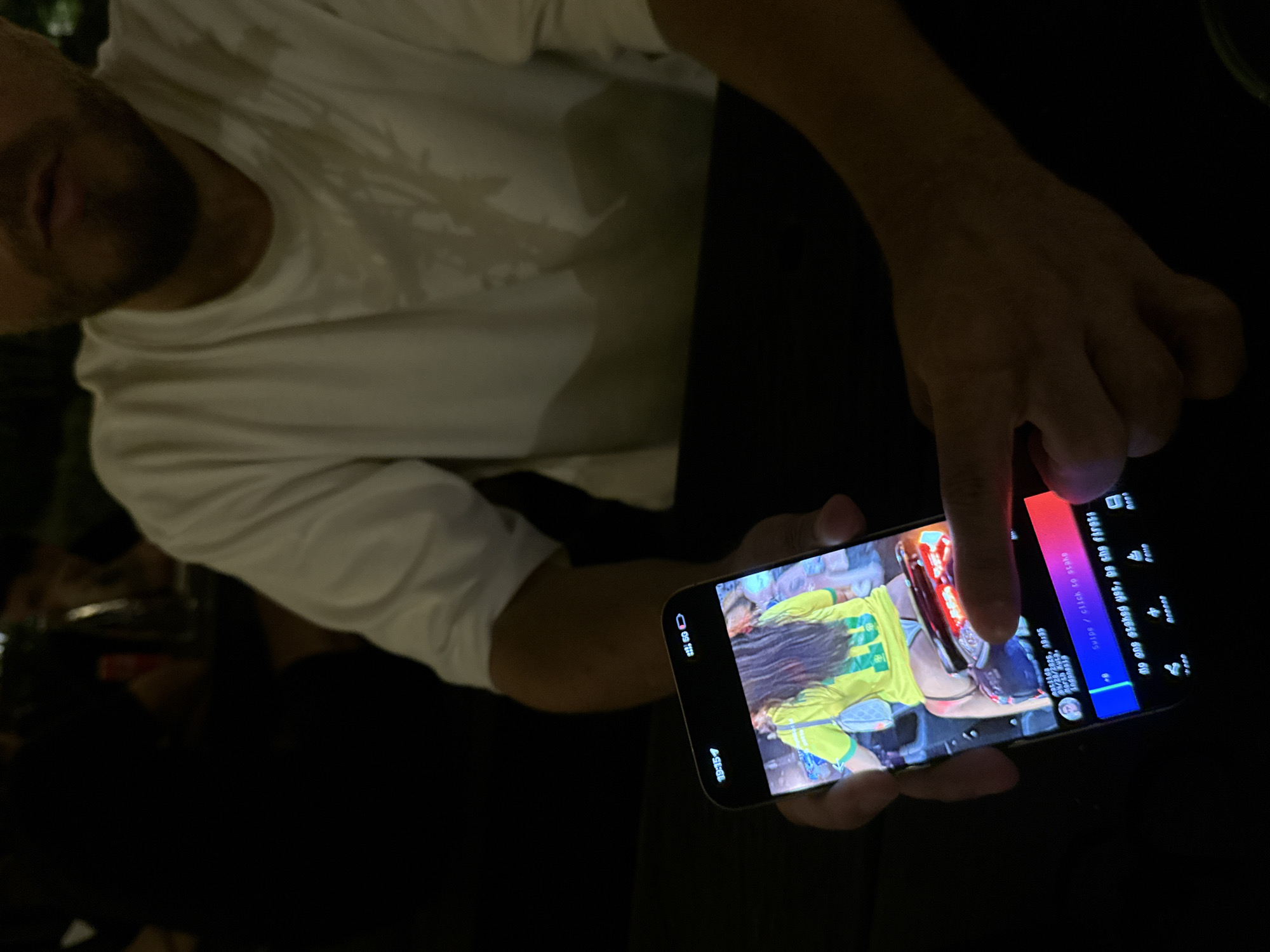 The image captures a partial view of a person engaging with a smartphone in a dimly lit environment, likely indoors at night. The subject, a person wearing a white t-shirt, is visible from the chest up, with only a portion of their face, including the mouth and a beard or stubble, discernible. Their right hand holds the smartphone, with an index finger actively touching the screen.
The smartphone screen displays a vertical image of a person with long dark hair, wearing a bright yellow and green shirt, possibly a jersey, with a hint of a motorcycle or scooter beside them. The phone's interface shows several details: the current time "19:15," a battery indicator at "60%," and a username "matilda." Associated with the image are a date and time "04/28/2025, 10:35," and location tags "north futa. Indonesia." The interface also presents interactive options like "Swipe / click to stake" and a social prompt stating, "No one staked yet, be the first!" A number "84" is visible, possibly indicating a count of stakes or engagement. The scene conveys a moment of casual digital interaction.