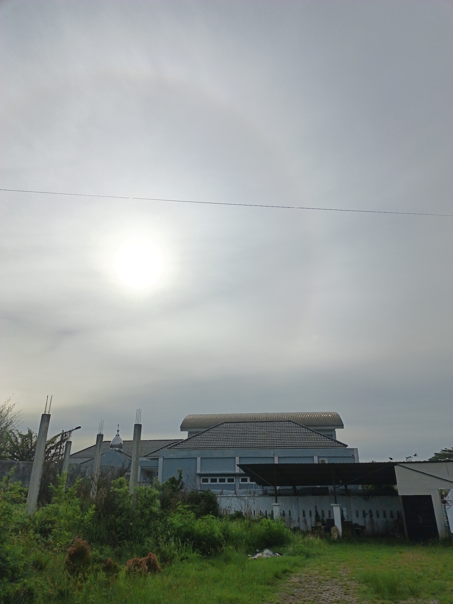 The image shows a cloudy sky with the sun obscured by the haze, a single overhead wire stretches across the sky. Below the sky, there is a large light blue building with a grey tiled roof. The building appears to be a mosque with a dome visible behind the building. A large awning is attached to the side of the building, covering what may be an outdoor gathering space. A white wall runs along the property line, and overgrown vegetation, including tall grass and shrubs, obscures much of the area between the building and the camera. The location is Lhokseumawe, Indonesia.