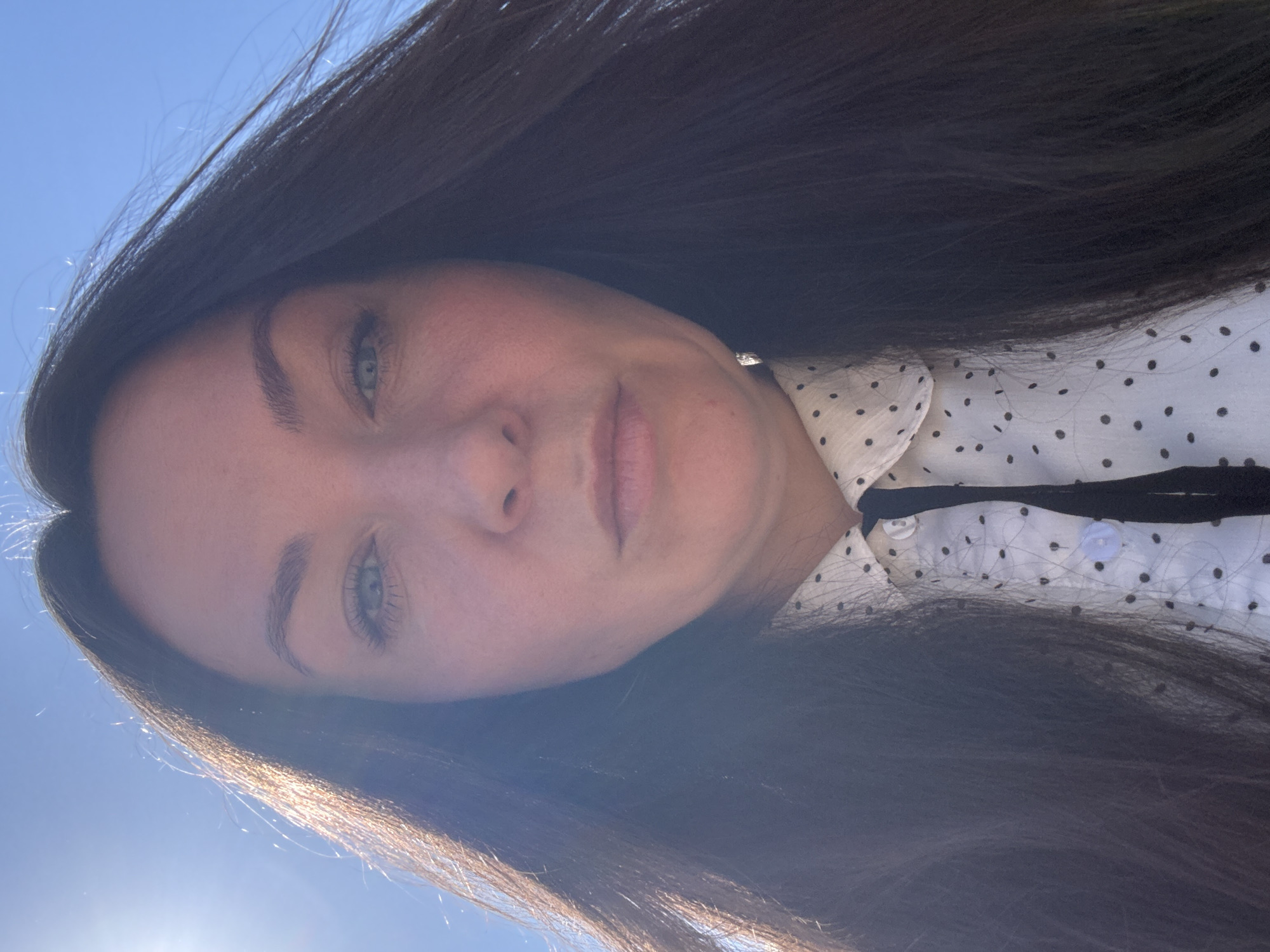 A woman with dark hair is pictured from a low angle, likely taken outdoors in bright sunlight. She is wearing a white blouse with black polka dots. The sky is visible in the background. The location is not discernible as being in the city of Фоки, Russia.