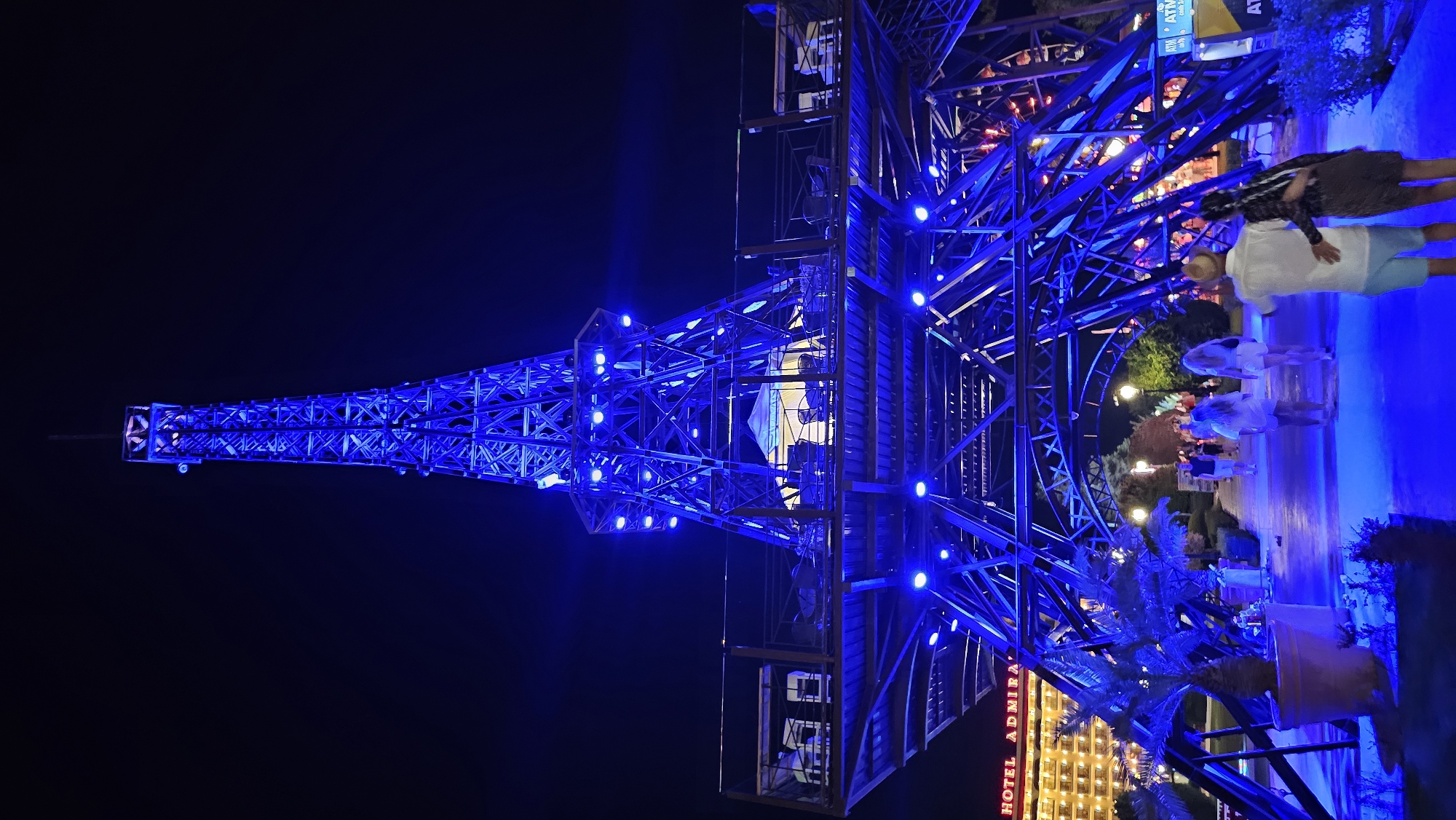 This is a nighttime, wide-angle, vertical shot of a replica Eiffel Tower, illuminated with vibrant blue lights. The tower's intricate metal latticework is highlighted by numerous blue LED lights, creating a striking contrast against the dark night sky.
In the foreground, the lower sections of the tower structure are visible, with some people standing nearby. On the right side of the image, three individuals are visible, two of whom appear to be a couple embracing, while another person stands slightly apart. Further into the background, other individuals are seen walking along a path. A palm tree in a large pot is also in the foreground, bathed in the blue light.
Behind the tower, a hotel building with illuminated windows is visible. There is also text visible on a sign that reads "HOTEL ADMIRAL". The overall scene appears to be a recreational area or a resort, likely a tourist attraction given the presence of a recognizable landmark replica. The blue lighting suggests a festive or decorative atmosphere. The image is captured at night, and the weather appears clear. The location is Varna, Bulgaria.