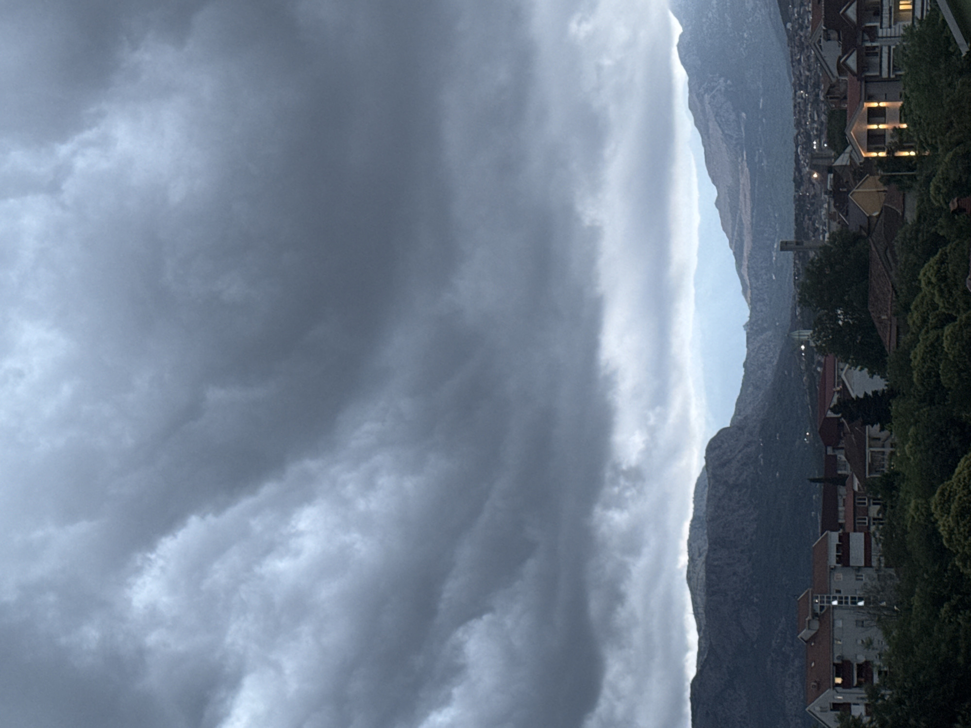 The image displays a panoramic view of a city nestled beneath a rugged mountain range, under a dramatically clouded sky. The vast majority of the sky is covered by dense, dark gray clouds, suggesting heavy overcast conditions, possibly indicating an impending storm or recent rainfall. A lighter strip of pale sky is visible between the cloud cover and the distant mountain peaks.
A large, imposing mountain range forms the central backdrop, its slopes appearing dark and rocky, stretching across the frame. Below the mountains, an urban area is visible, primarily on the right side, featuring numerous buildings with varying architectural styles and predominantly reddish-brown roofs. Several of these buildings show illuminated windows and outdoor lights, indicating either late evening or extremely dim, overcast daytime conditions. Dense green trees and foliage are present, particularly on the right, partially obscuring some structures and adding natural elements to the cityscape.
The scene captures a quiet moment with no direct human activity visible. The overall impression is one of subdued light and a potentially moody atmosphere due to the heavy cloud cover. The location context provided is Podgorica, Montenegro. There is no visible text in the image.