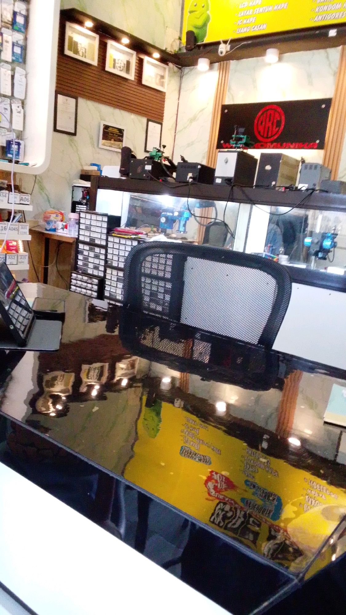 The image depicts the interior of a small electronics repair shop, likely in Cibenda, Indonesia, judging by the style and some visible text. The shop appears to be well-lit and organized, with various electronic components and equipment visible.
In the foreground, a large, glossy black table dominates the scene, reflecting the objects above it. On the table, a laptop or tablet is propped up, its screen displaying some text or interface. To the right of the table, a black office chair with a mesh back is positioned.
Behind the table, shelves are filled with small drawers or containers, likely holding electronic parts. Various electronic devices, possibly repair equipment like soldering stations or power supplies, are arranged on shelves and a counter. A prominent black sign with a red circular logo and text that appears to be "ARC RUNNING" is mounted on a wooden-paneled wall. Above this sign, a yellow banner with text in what appears to be Indonesian, along with a cartoonish green character, is visible.
On the walls, several framed pictures or certificates are hung, adding a touch of decoration. The overall impression is of a functional retail and repair space, dedicated to electronics services. The environment is clean and orderly, suggesting a professional operation.
