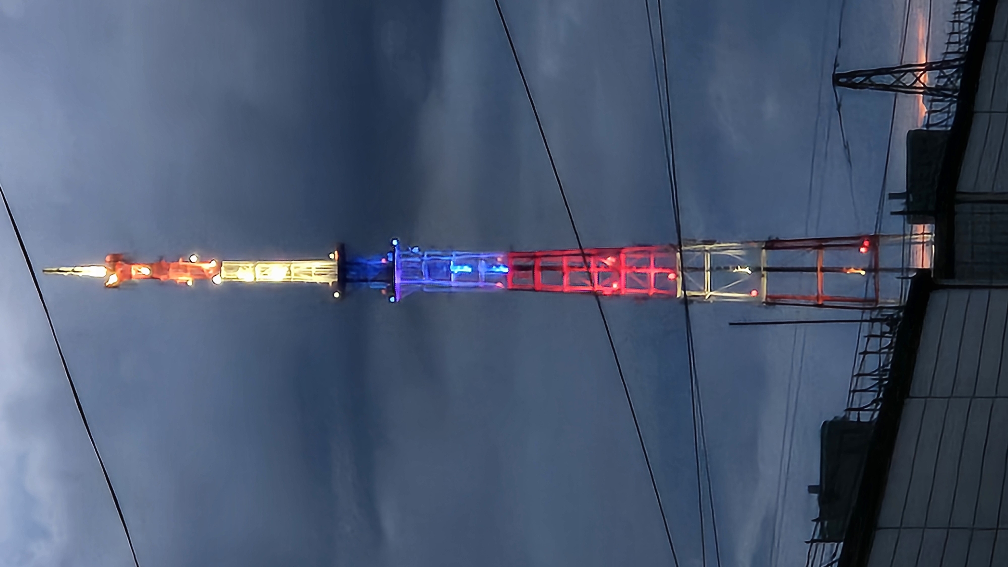 A tall, lattice tower, illuminated with red, white, and blue lights, stands against a dark, cloudy sky. The tower is segmented into sections, with the lower portion lit in red, the middle in blue, and the upper sections in white and red. A pointed antenna structure tops the tower. In the foreground, the tops of buildings and electrical wires are visible. The overall impression is that of an urban landscape at dusk or during a cloudy day, with the tower serving as a prominent, brightly lit landmark.