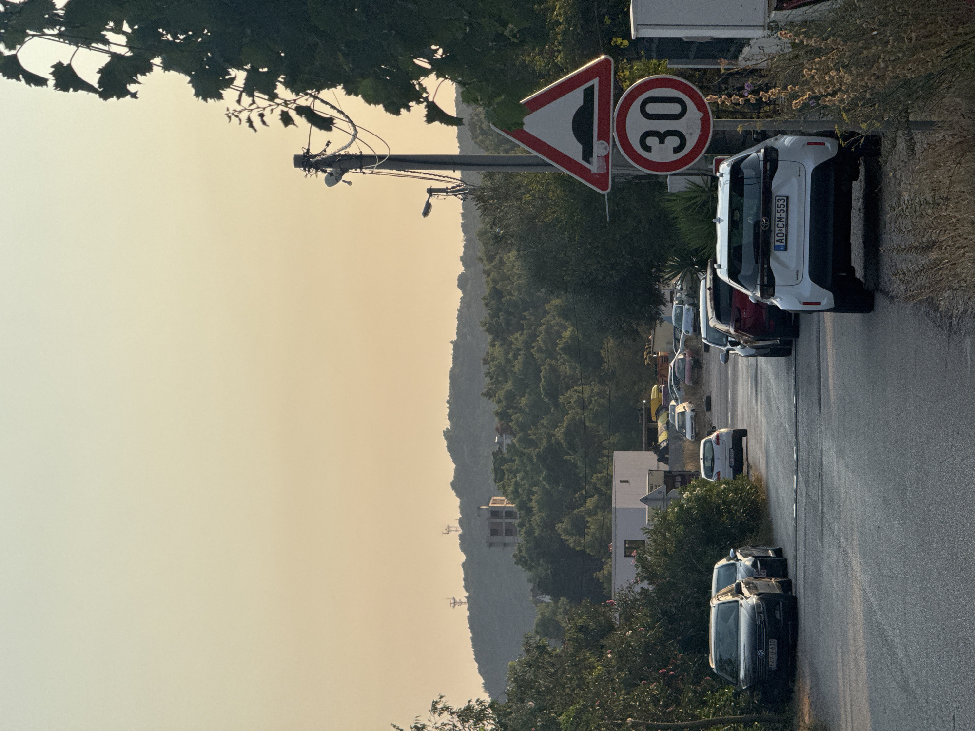 The image depicts a street scene in Sutomore, Montenegro, during what appears to be late afternoon or early evening, given the warm, diffused light. The road is asphalt and shows some wear. On the right side of the road, a utility pole stands with several signs attached. The topmost sign is a triangular warning sign with a red border and a black graphic depicting a speed bump. Below it is a circular speed limit sign with a red border and a white background, displaying the number "30" in black. A white Toyota Yaris is parked on the right side of the road, immediately behind the speed limit sign, with its license plate reading "AO CM 553". Further down the road, several other vehicles are parked and driving.
On the left side of the road, a dark colored Volkswagen Passat is parked, with its license plate visible as "E47-O-610". Adjacent to it, another car is partially visible. In the background, a white building with visible windows is situated among dense green trees. Beyond the trees, rolling hills rise, with the silhouettes of at least two communication towers visible on the highest points. The sky above is a soft, gradient of pale yellow and orange, indicating the time of day. Vegetation, including bushes and tall grasses, lines the sides of the road and extends into the background. The overall atmosphere suggests a quiet, residential or semi-rural area of Sutomore.