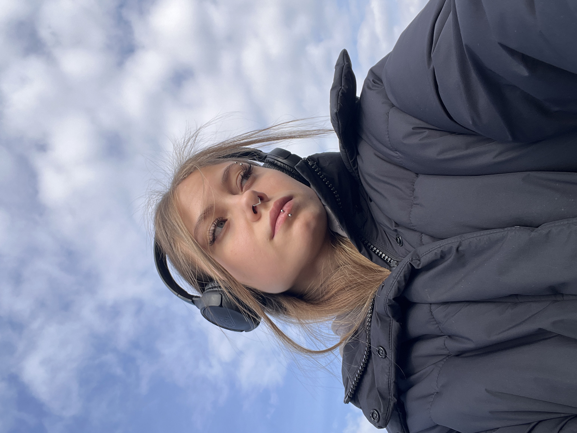 A young woman stands outdoors in Фоки, Russia, under a sky filled with scattered clouds. She wears a black padded jacket with a zipper and buttons. She has headphones around her head. She has blonde hair, and her face is angled towards the camera. She has piercings on her eyebrow, nose, and lower lip.